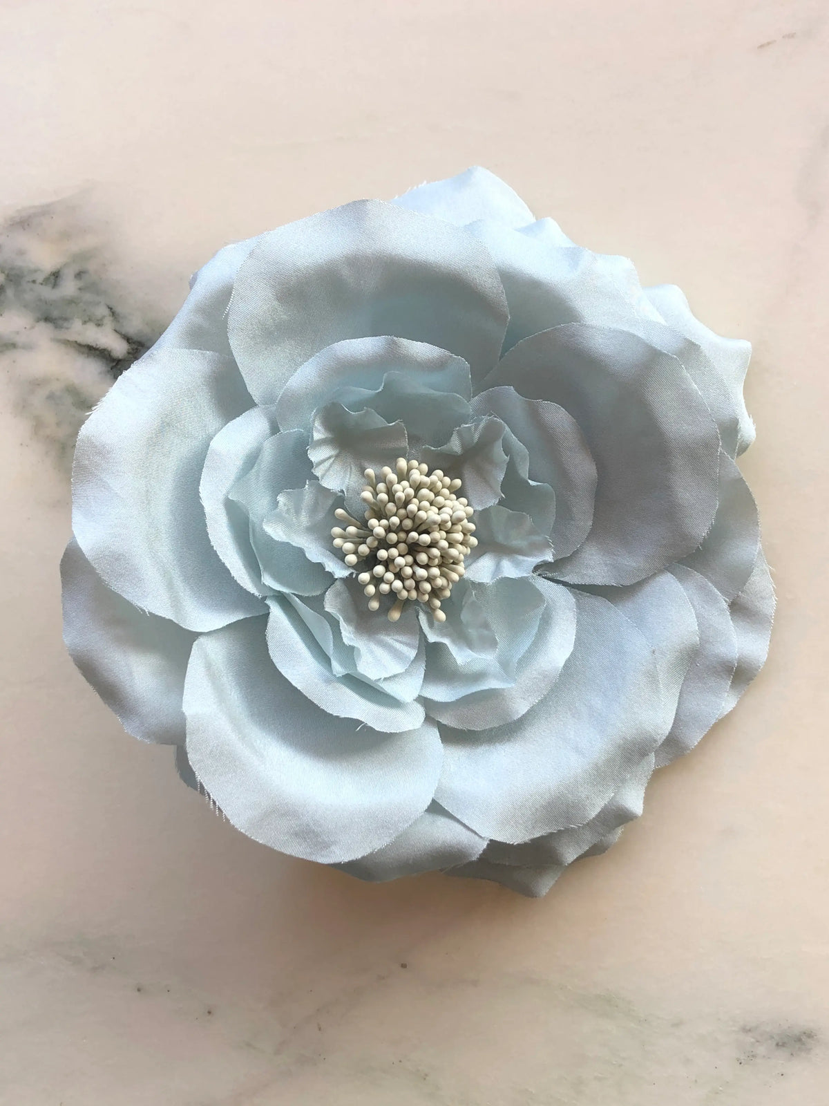 Broochella Men's Signorina Fioritina Flower Brooch Pin in sky blue with layered fabric petals and a cream stamen centre, photographed on a white marble background with grey veining.
