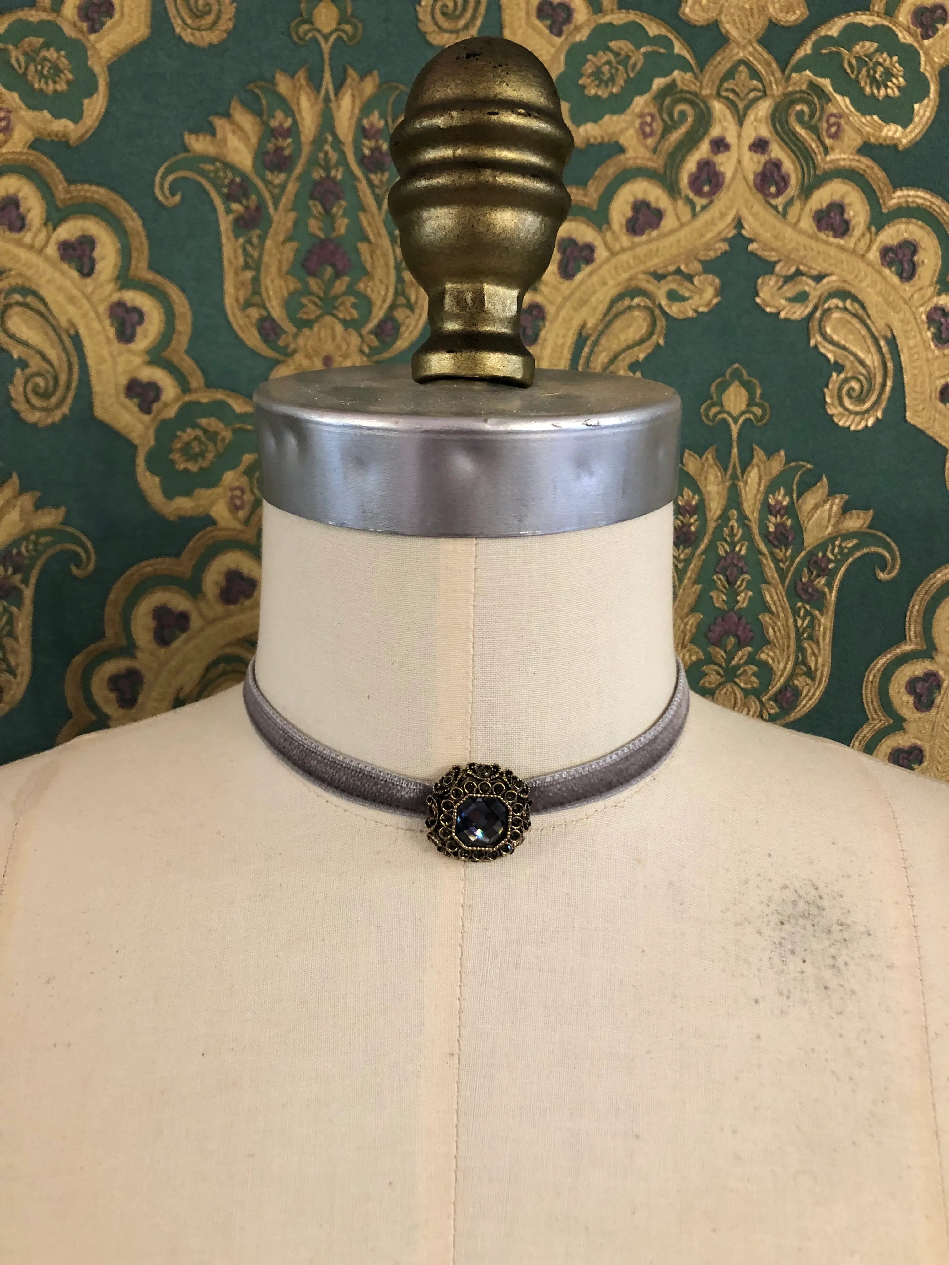 A Broochella Lady Whistledown silver velvet choker necklace with a decorative antique bronze filigree centrepiece, displayed on a cream-coloured dress form against a patterned teal and gold damask wallpaper background.