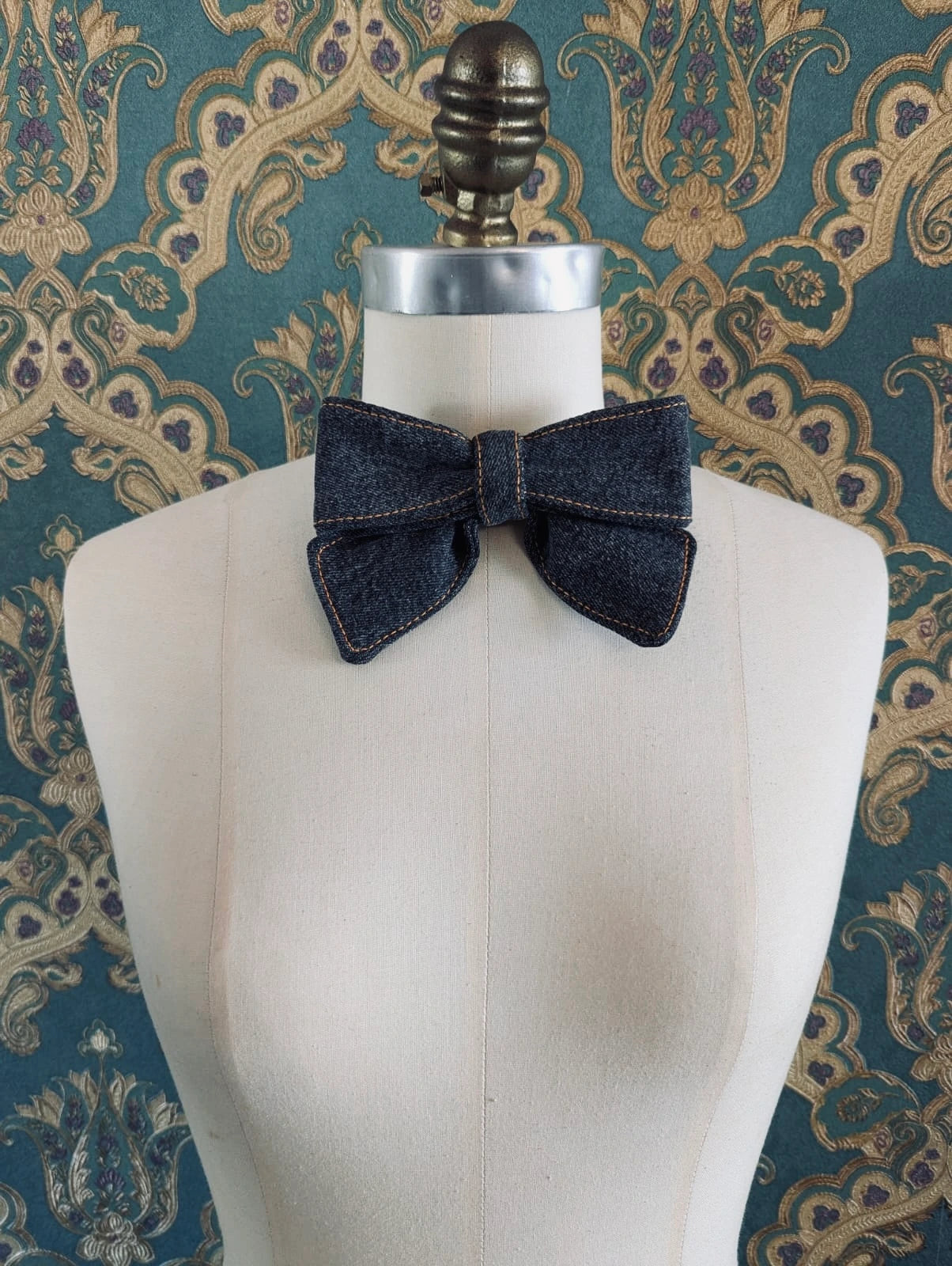 Broochella handmade black small denim bow brooch. Up-cycled form vintage denim jeans. It is pinned to a mannequin. The background is green and gold damask wallpaper. 