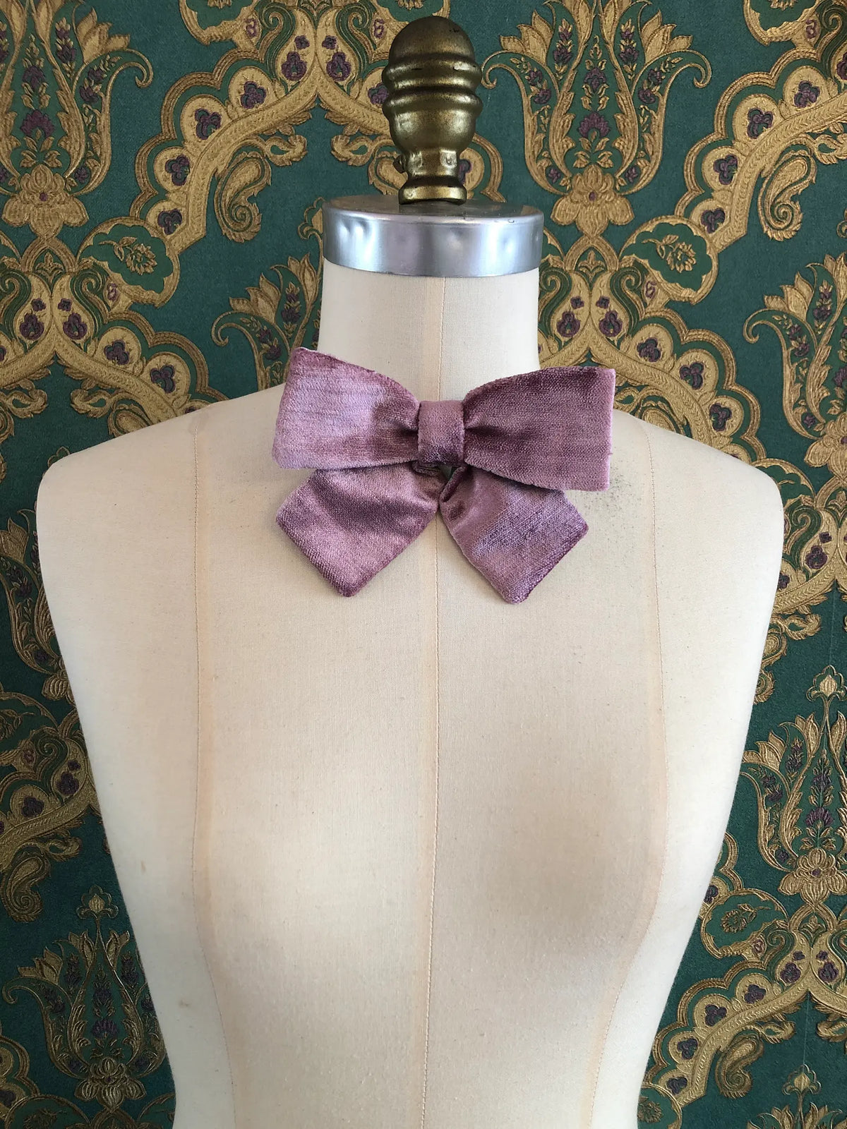 Broochella handmade Small Velvet Bow Brooch in soft violet velvet with contrasting green lining, styled on the shoulder of a cream mannequin against ornate green and gold wallpaper.
