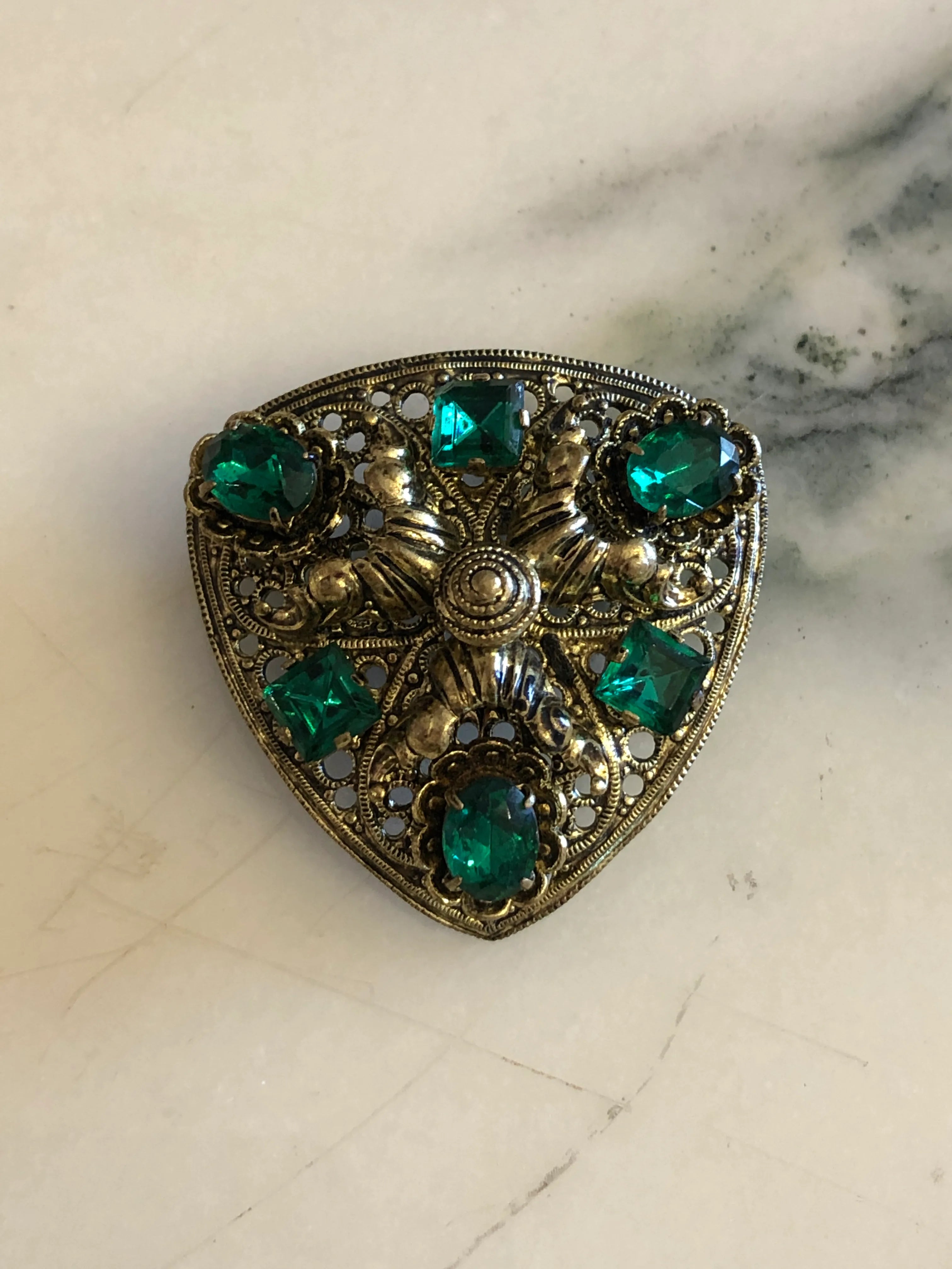 Broochella Smaraldo vintage Czech shield brooch with emerald-green crystals and ornate antiqued silver-tone filigree. the background is white marble