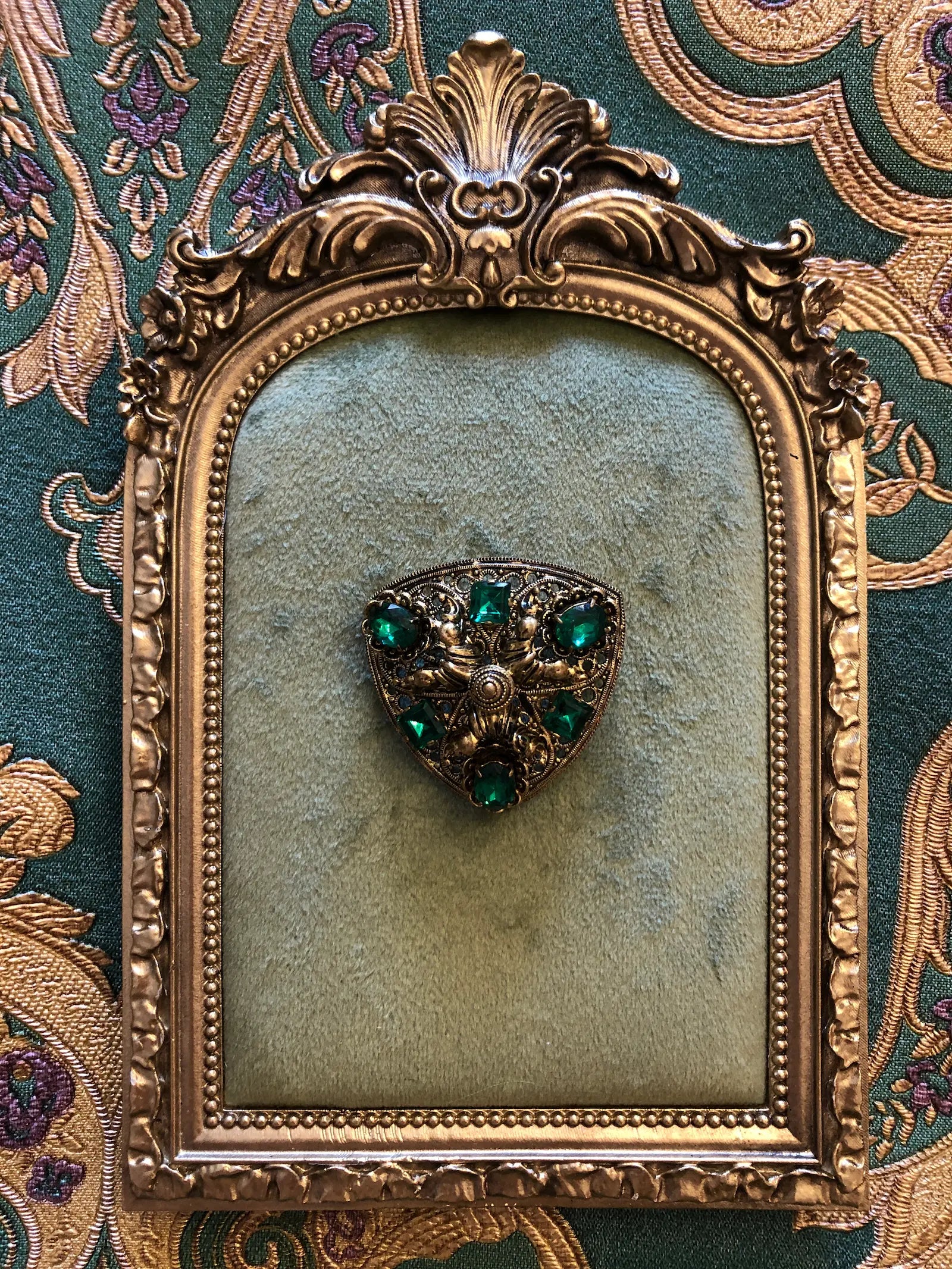 An ornate gold decorative frame with a Broochella Smeraldo Vintage Czech shield-shaped brooch featuring green gemstones, set against a patterned green and gold fabric background.