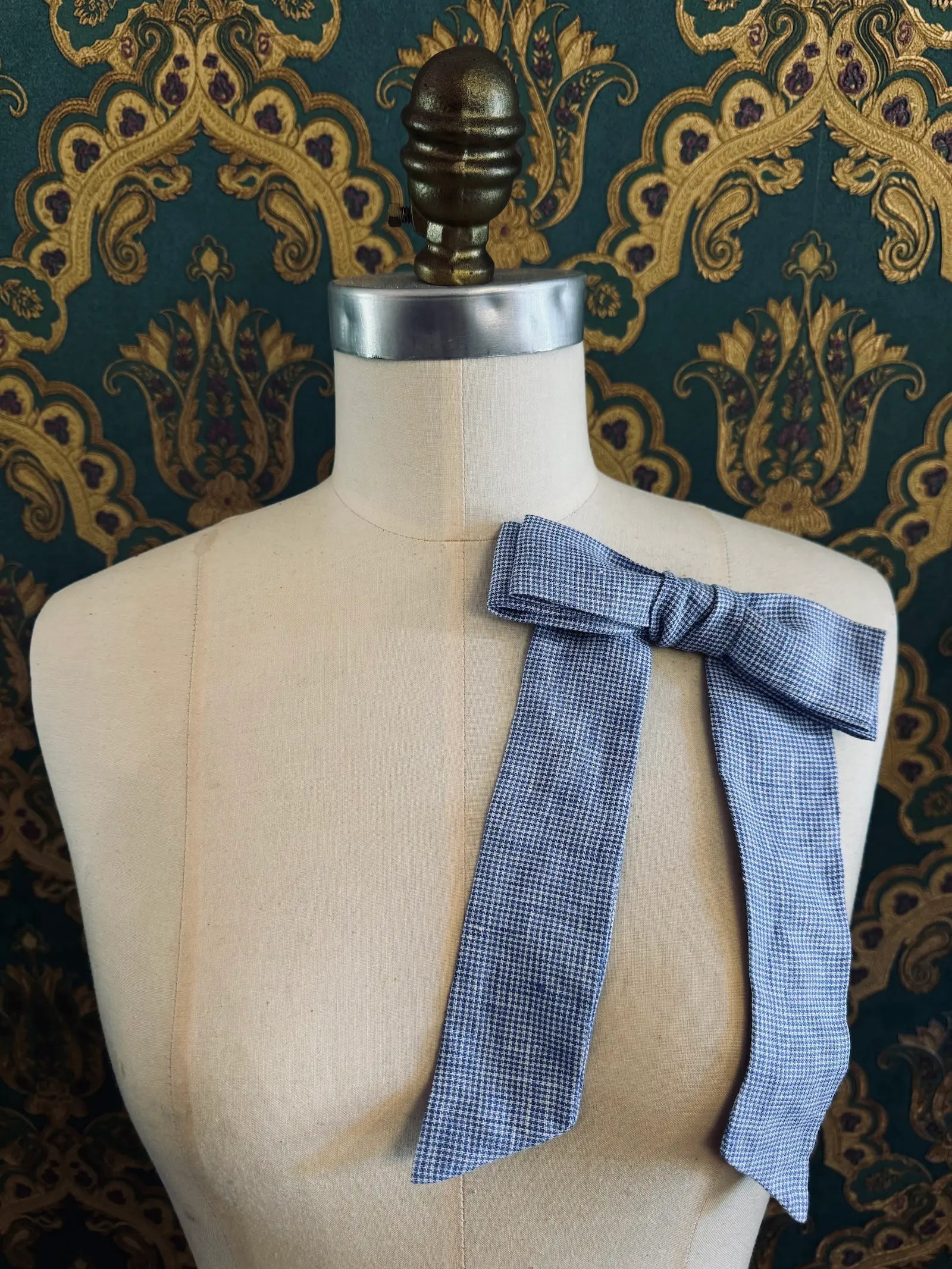 Broochella  Sofia handmade linen bow brooch in blue check. Pictured on a mannequin. The background is green and gold damask wallpaper.