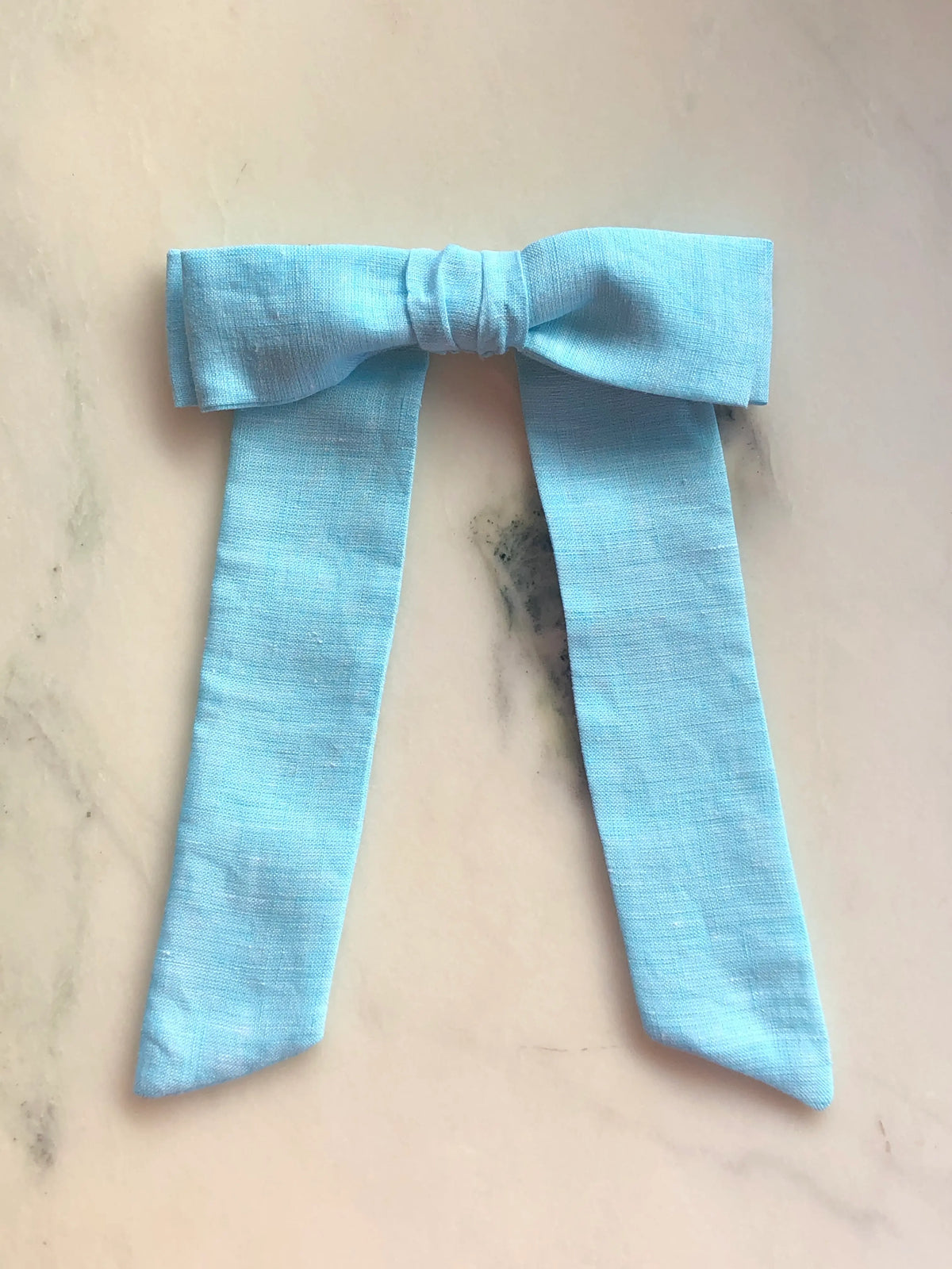 Broochella Men's Sofia blue linen bow brooch, a handmade designer statement accessory with an oversized bow and elegant long ribbon tails, photographed on white marble.