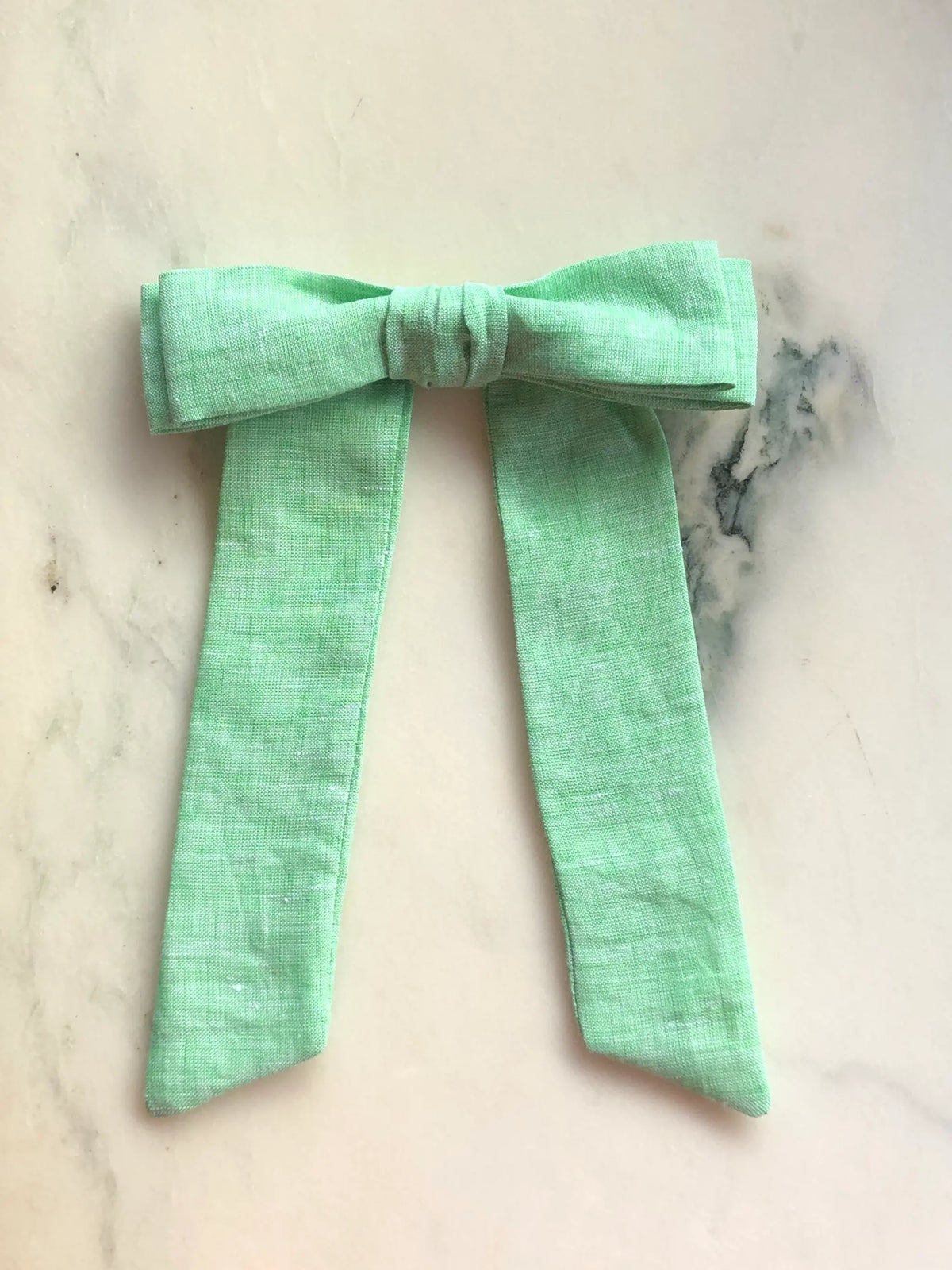 Broochella Men's Sofia green linen bow brooch, a handmade designer statement accessory with an oversized bow and elegant long ribbon tails, photographed on white marble.