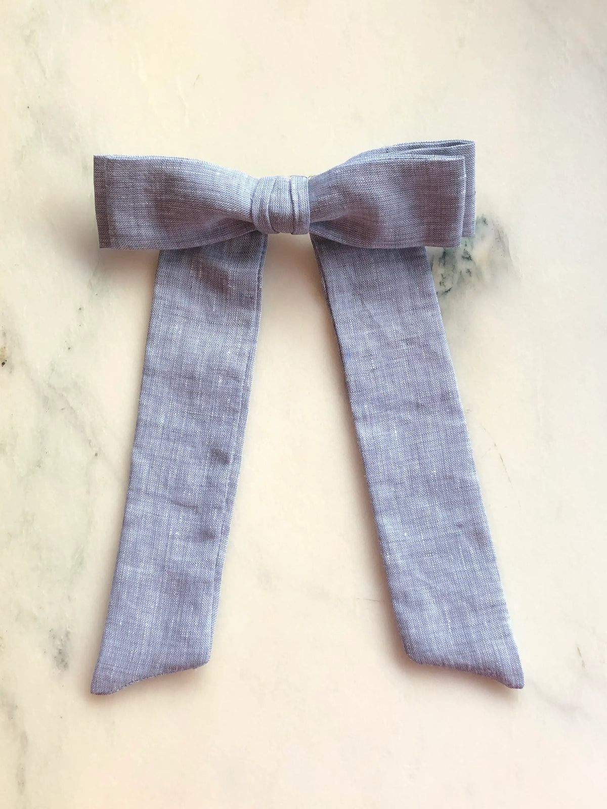 Broochella Men's Sofia grey linen bow brooch, a handmade designer statement accessory with an oversized bow and elegant long ribbon tails, photographed on white marble.