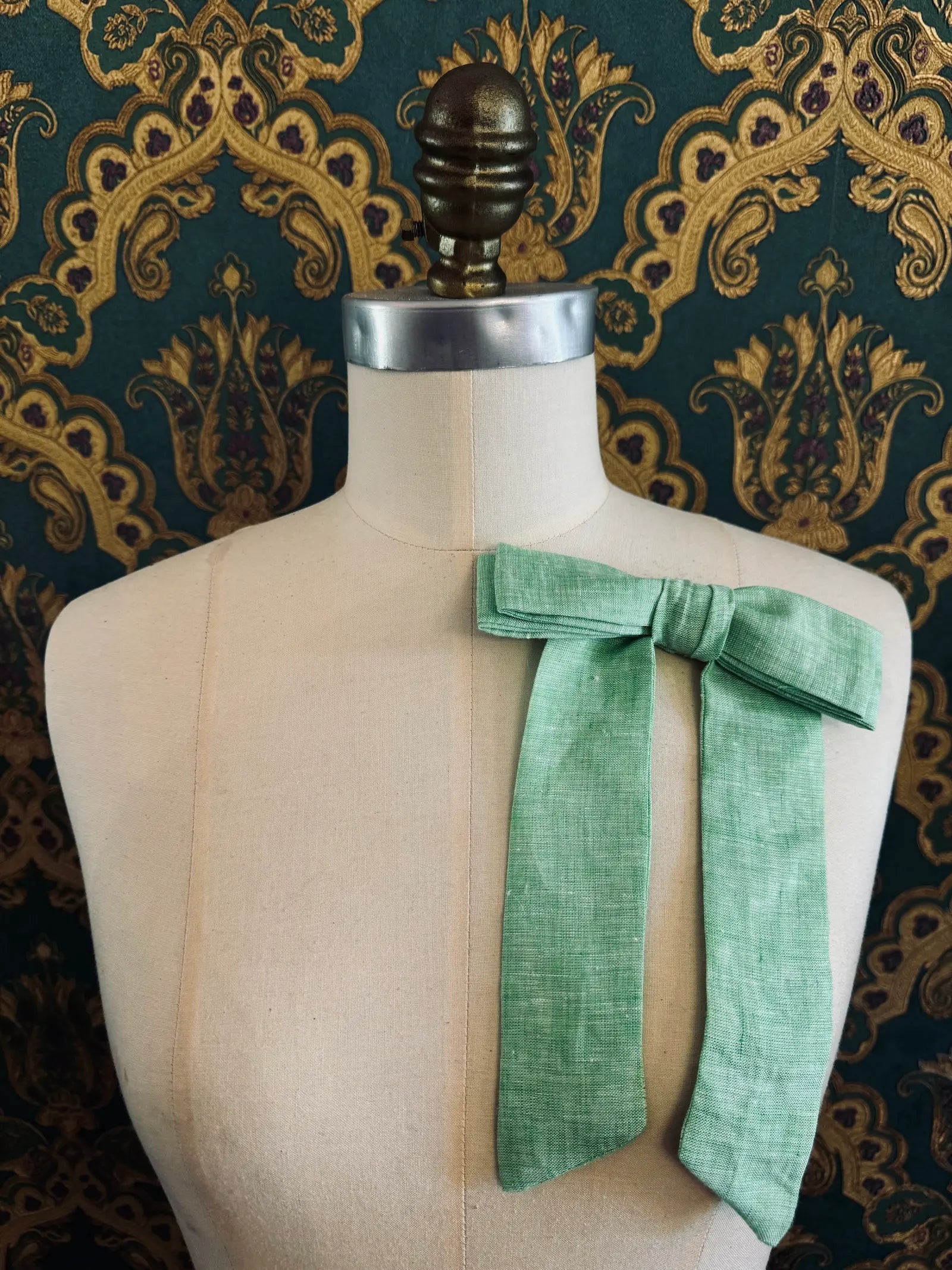 Broochella  Sofia handmade linen bow brooch in lime. Pictured on a mannequin. The background is green and gold damask wallpaper.