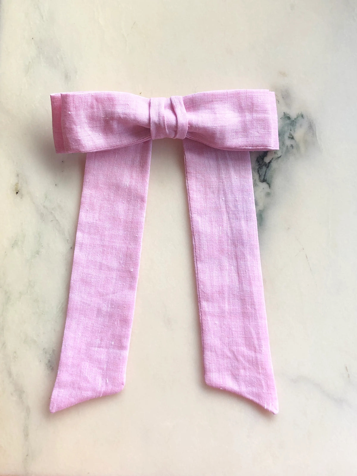 Broochella Men's Sofia pink linen bow brooch, a handmade designer statement accessory with an oversized bow and elegant long ribbon tails, photographed on white marble.