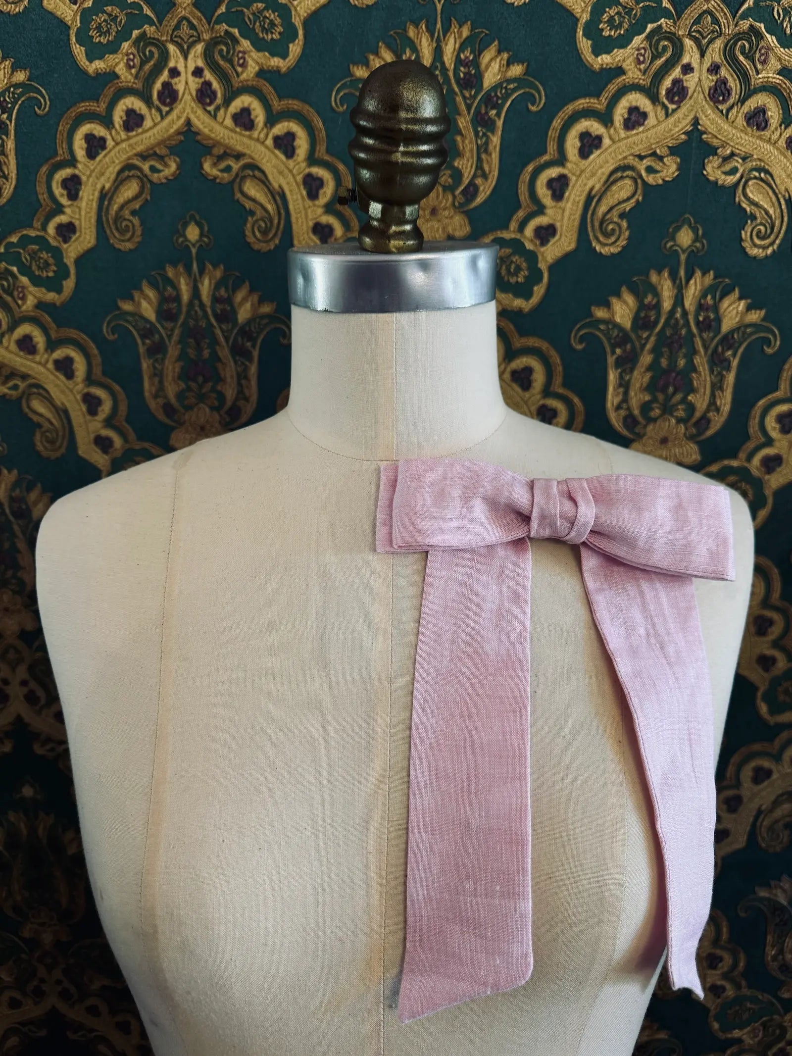 Broochella  Sofia handmade linen bow brooch in pink. Pictured on a mannequin. The background is green and gold damask wallpaper.