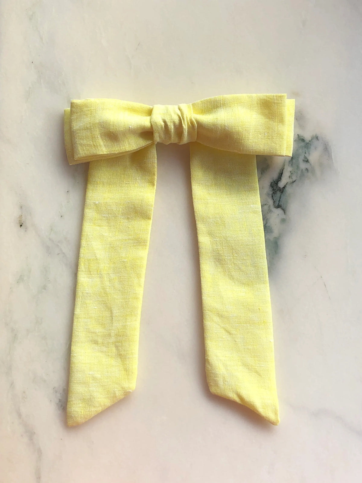 Broochella Men's Sofia yellow linen bow brooch, a handmade designer statement accessory with an oversized bow and elegant long ribbon tails, photographed on white marble.