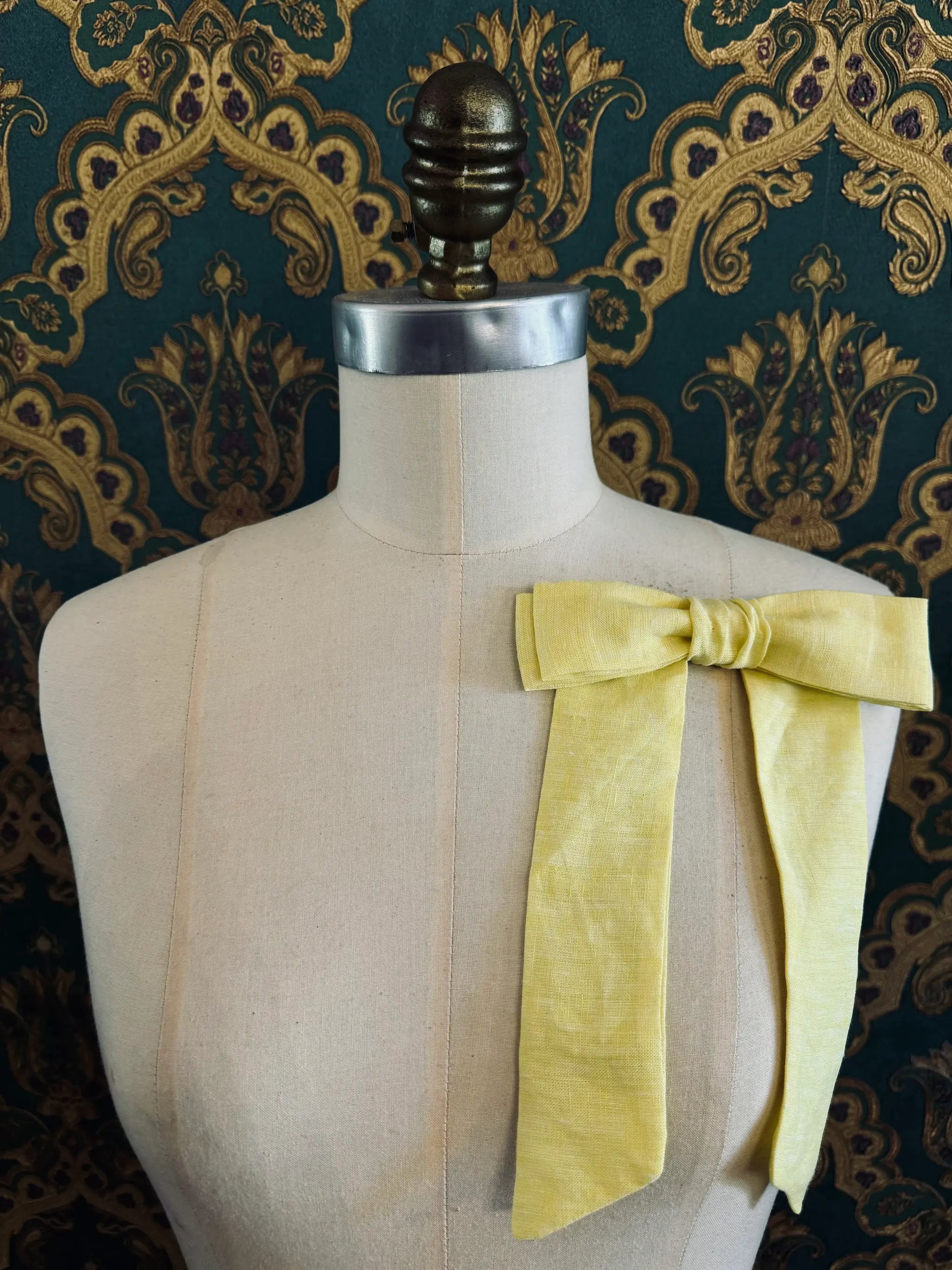 Broochella  Sofia handmade linen bow brooch in yellow. Pictured on a mannequin. The background is green and gold damask wallpaper.