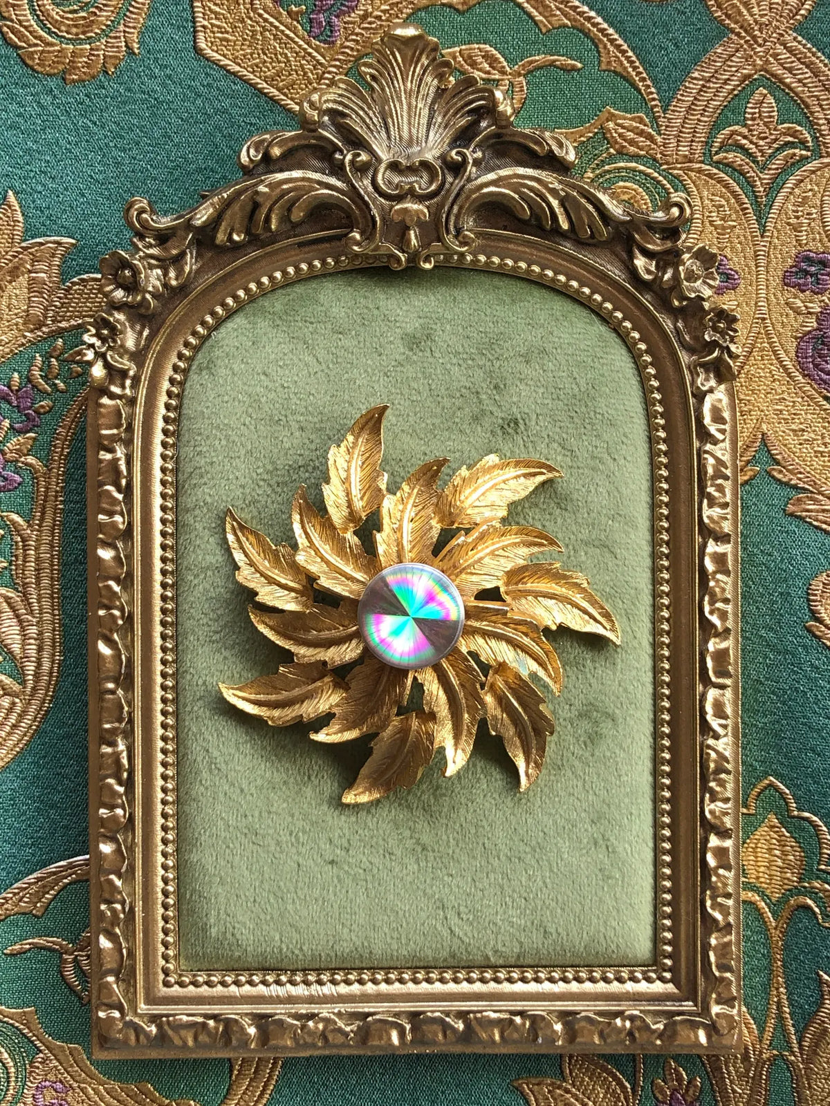 Broochella Sole d’Oro Vintage Brooch featuring sculptural gold-tone leaves and an iridescent rainbow-effect centre, displayed on sage green velvet in an ornate antique-style gold frame against green and gold patterned fabric.