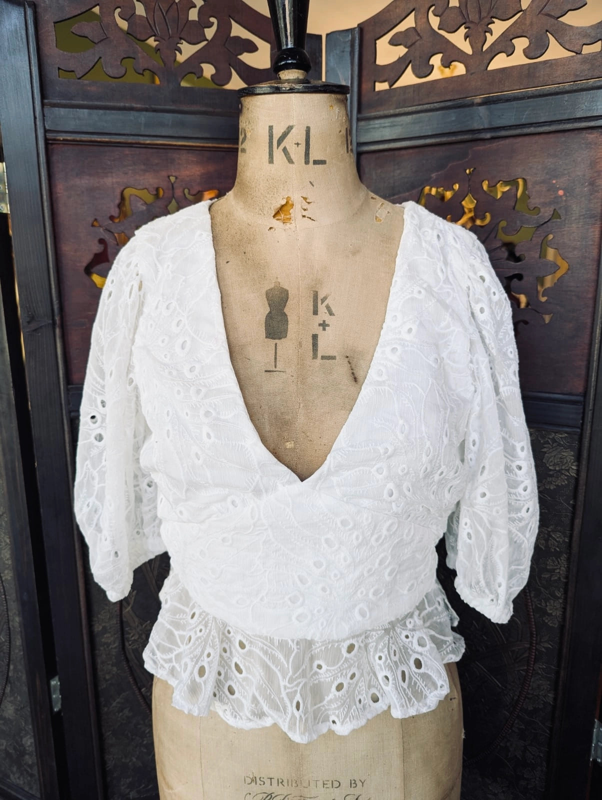 Broochella Song of style white size m white cotton broderie eyelet blouse with peplum hem and v neck. It is pictured on a mannequin The background is and ornate dark wood room divider. 