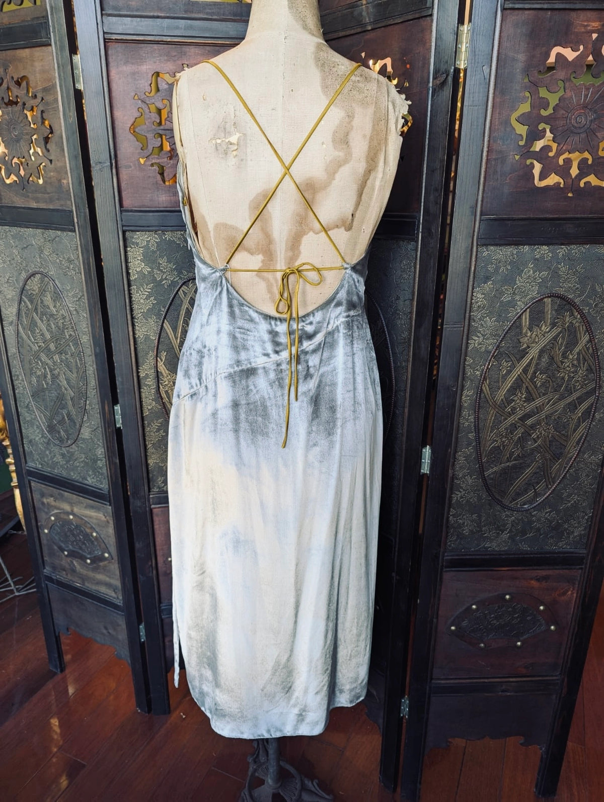 The back view of Broochella Ssone silver velvet slip dress, with gold straps. It is pictured on a mannequin. The background is an ornate, dark wood room divider. 