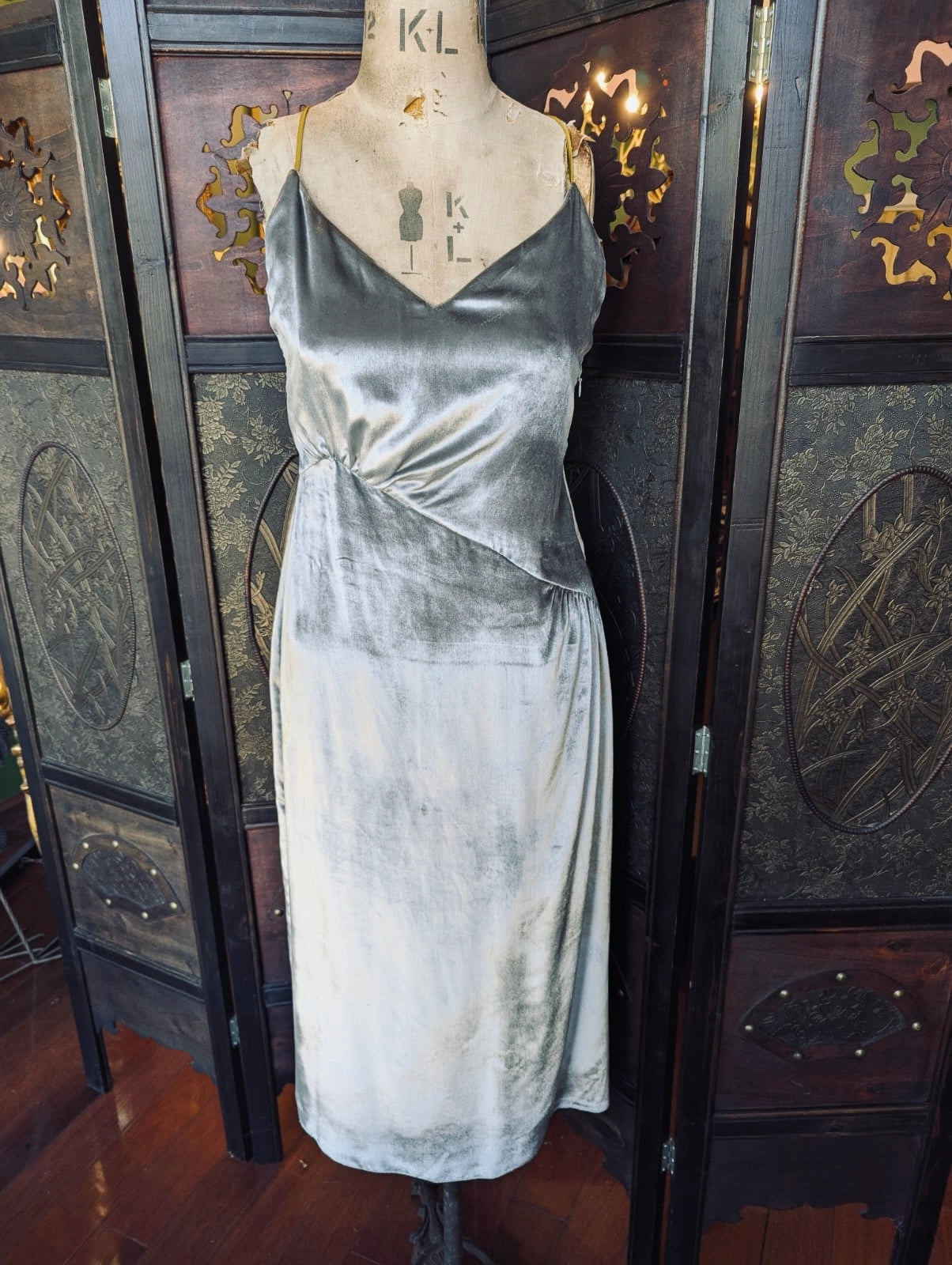 Broochella Ssone silver velvet slip dress, with gold straps. It is pictured on a mannequin. The background is an ornate, dark wood room divider. 