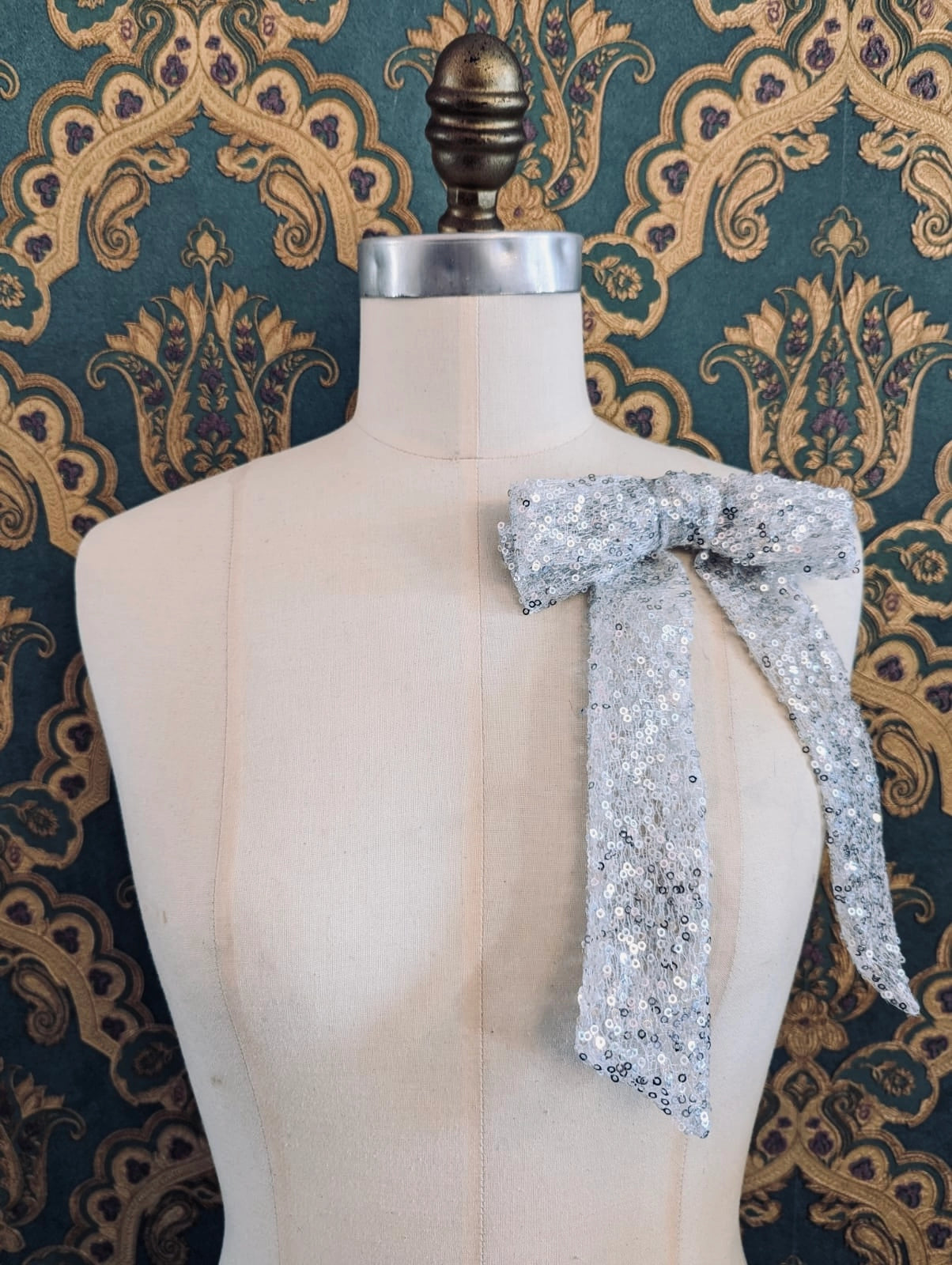 Broochella stardust handmade silver sequin bow brooch. The brooch is pinned to a mannequin at the neck. The background is green and gold damask wallpaper.