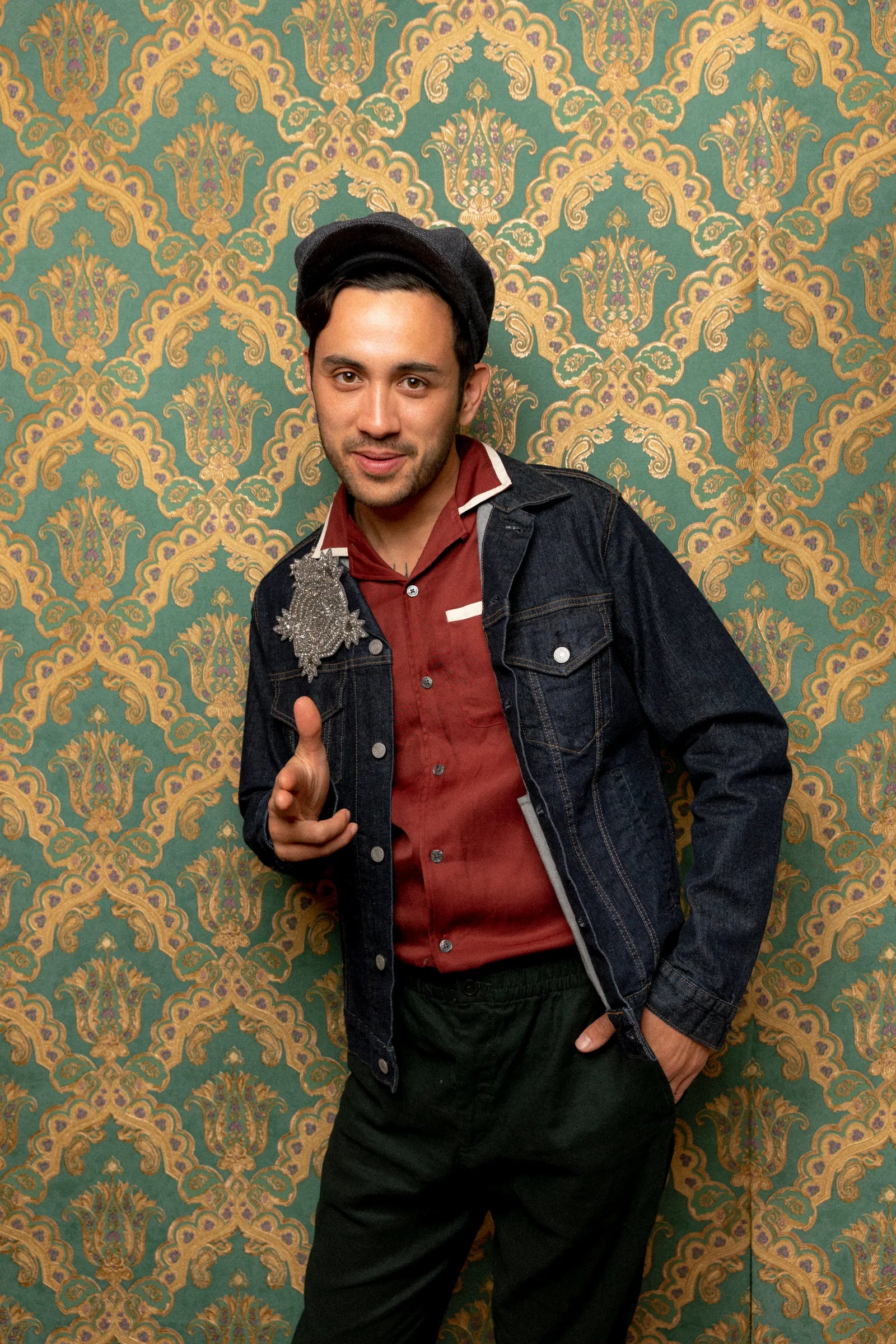 Male model with dark hair wearing a denim jacket and the Broochella Stella brooch. The background is green and gold damask wallpaper
