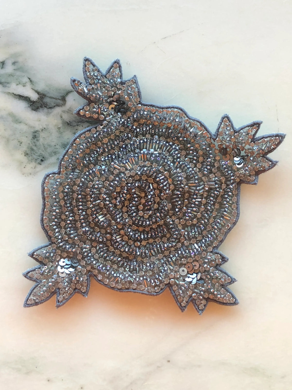 Broochella Stella Men's Hand-Beaded Brooch featuring an ornate floral design intricately embellished with silver, champagne and metallic beads, crystals and sequins on a cream marble background.