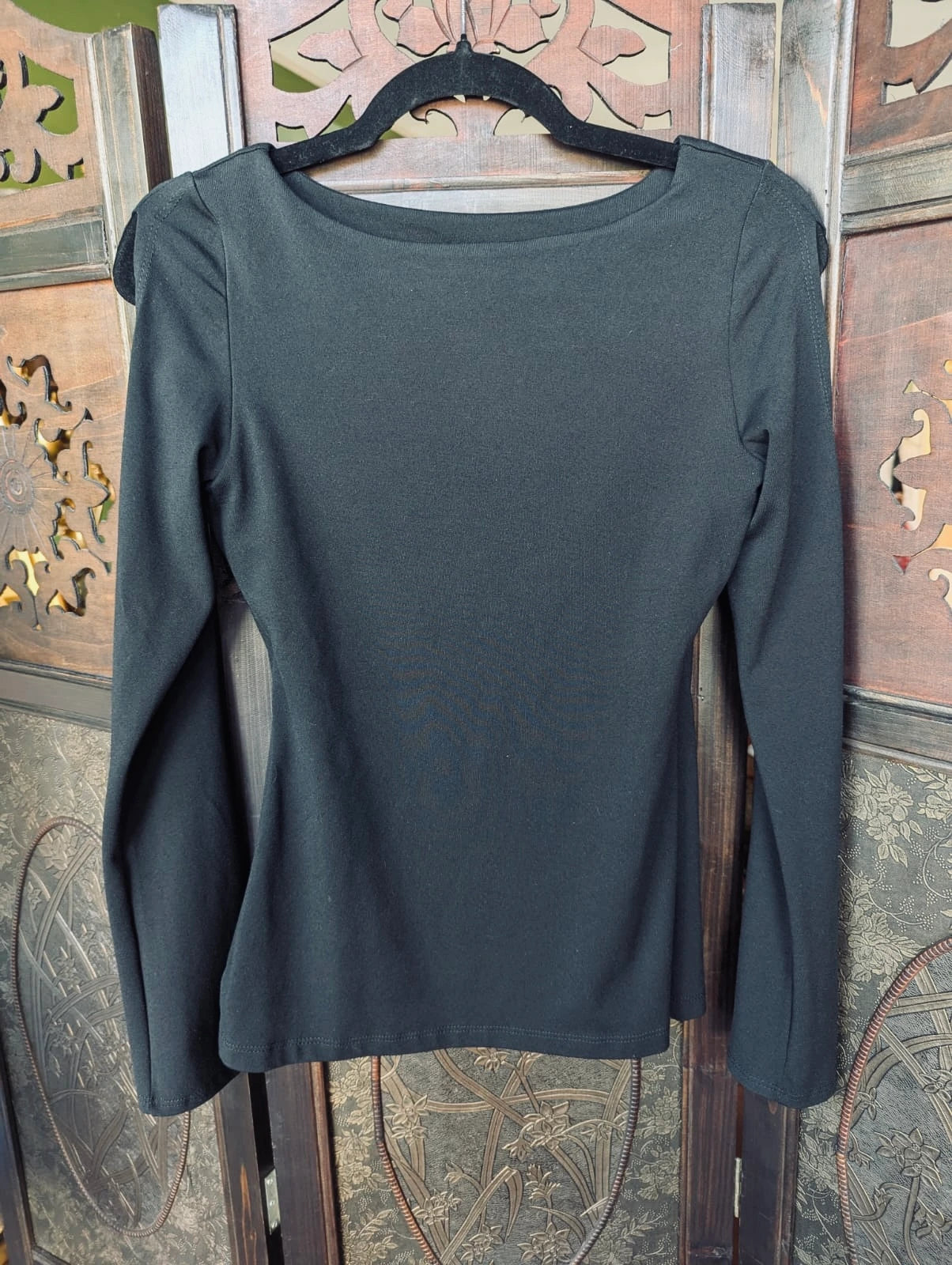 A Broochella Susana Monaco black  long-sleeve top with a boat neckline, hanging on a black hanger against a decorative wooden screen.