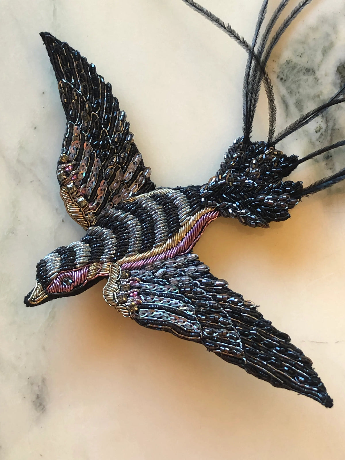 Broochella Swallow Bridal Bird Brooch featuring an intricately beaded black and metallic swallow in flight with sequinned wings and long feathered tail, photographed on a pale cream marble background.