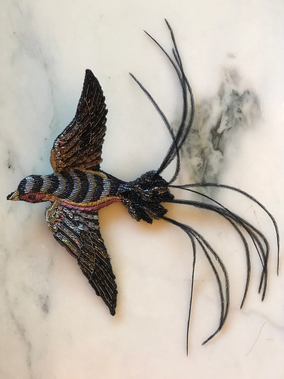 Broochella Swallow Bridal Bird Brooch featuring an intricately beaded black and metallic swallow in flight with sequinned wings and long feathered tail, photographed on a pale cream marble background.