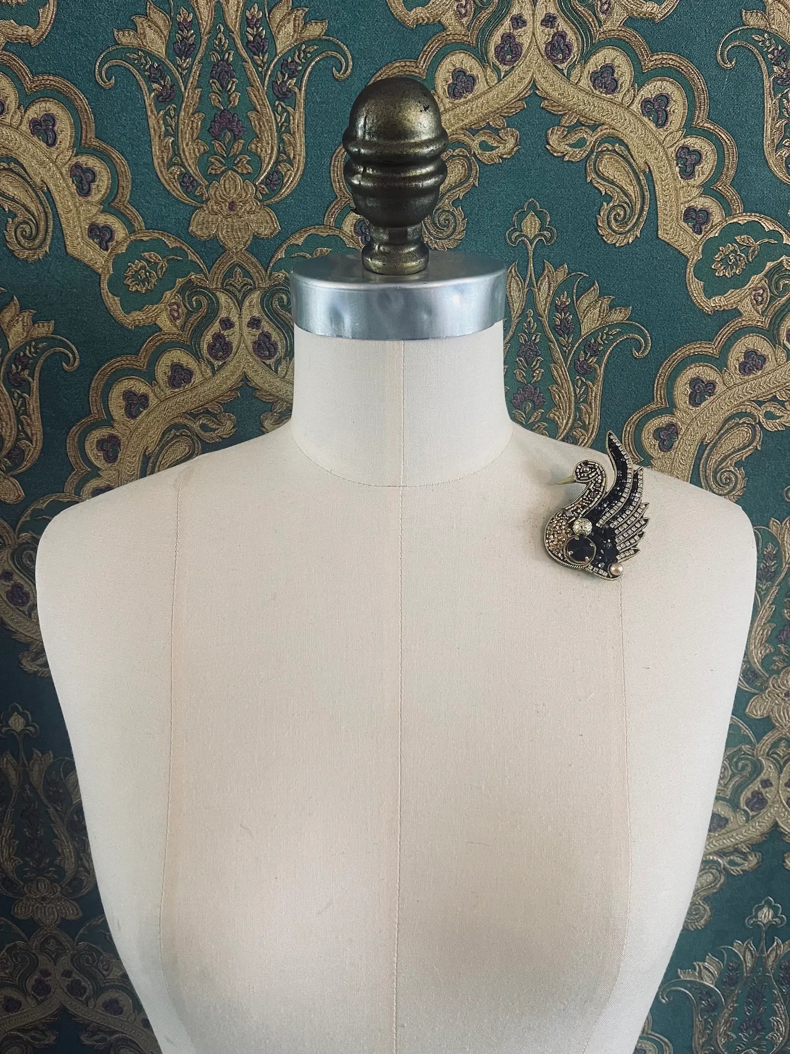 Broochella hand beaded black and gold figural swan brooch. Pictured on a mannequin. The background is green and gold damask wallpaper