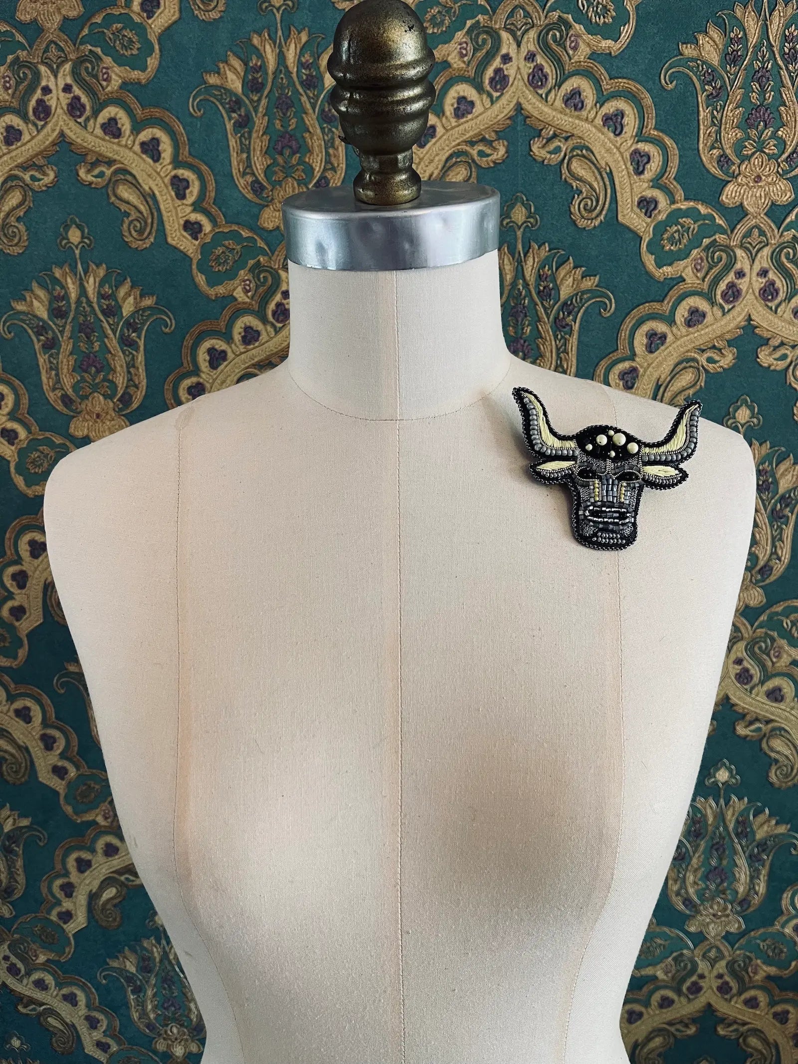 Broochella taurus hand beaded designer zodiac brooch. Styled on a mannequin. The background is green and gold damask wallpaper