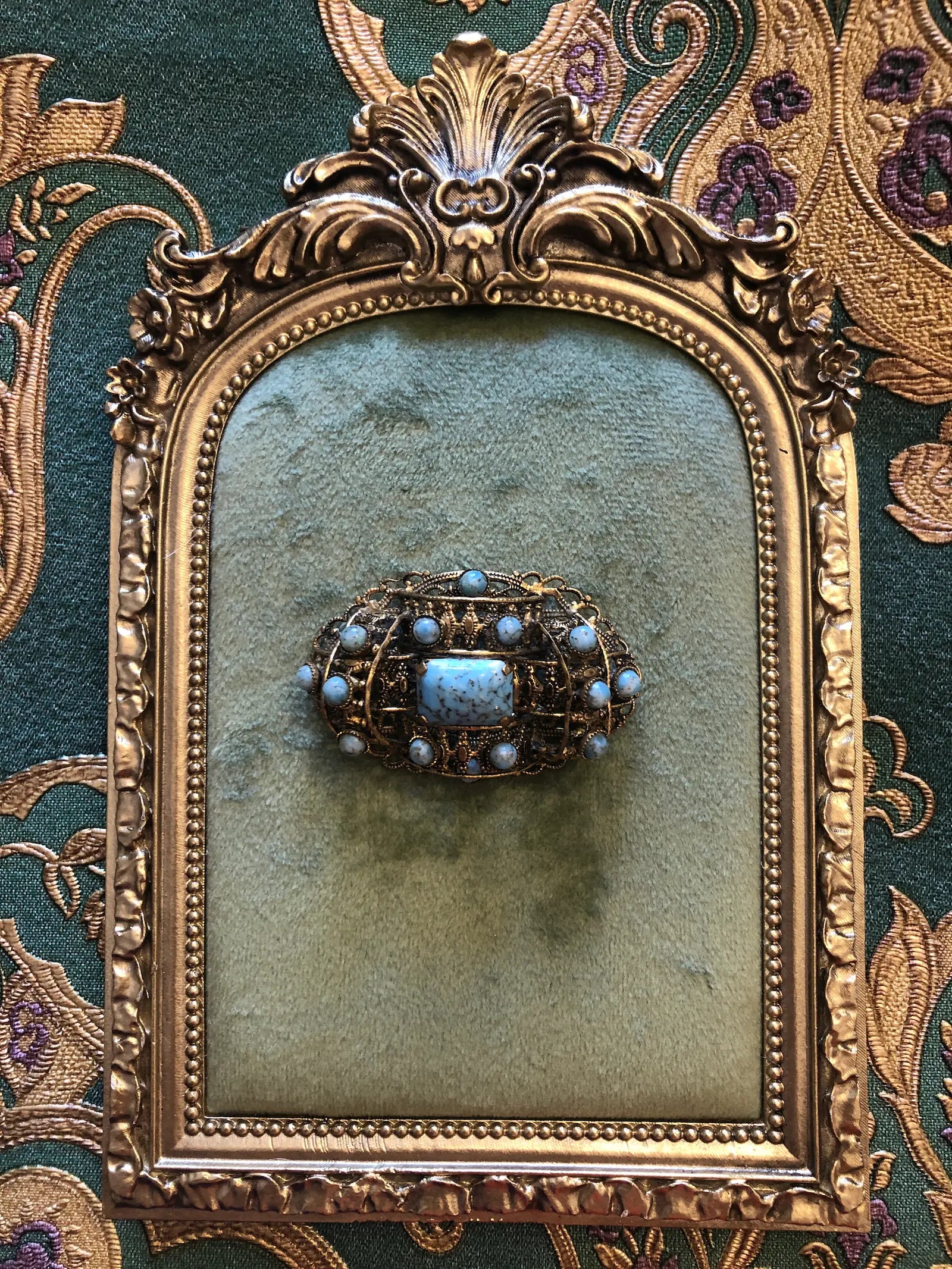 Broochella tesoro azzurro czech filigree vintage oval brooch with a turquoise-blue central stone and small light blue stones around it, set in gold-toned metal, displayed on a green velvet surface inside an antique gold frame against a patterned green and gold fabric background.