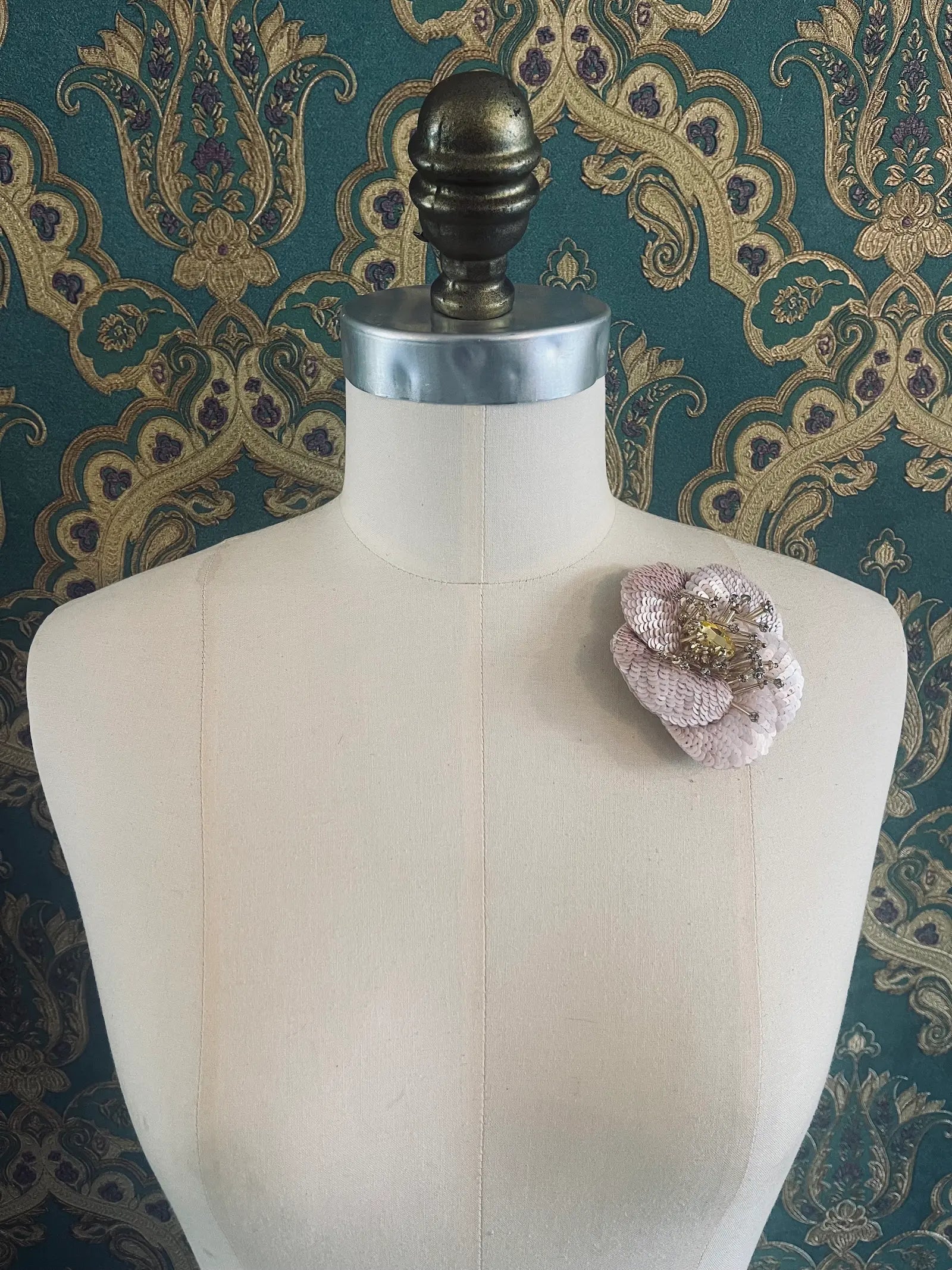 Broochella Tesoro ingioiellato pink and gold handmade sequin flower brooch. Pictured on a mannequin. The background is green and gold damask wallpaper