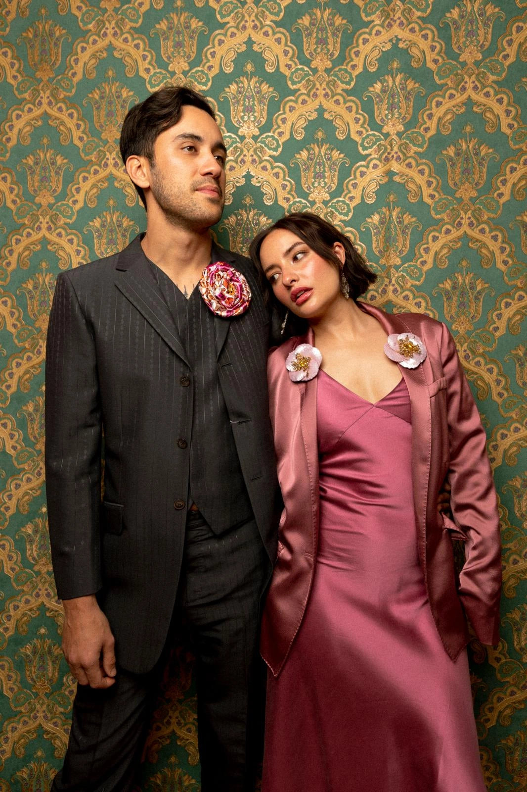 A man in a dark gray pinstripe suit with a pink floral boutonniere stands next to a woman in a dusty rose satin dress and matching jacket, posed against a green and gold damask wallpaper background. She is wearing two Broochella tesoro ingioiellato flower brooches.