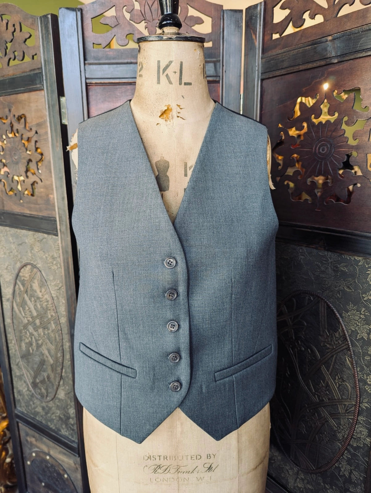 A Broochella Frankie Shop waistcoat vest in a muted slate blue-grey fabric, displayed on a vintage cream-coloured dress form mannequin. The vest features a button-front closure, notched lapels, and two front welt pockets.