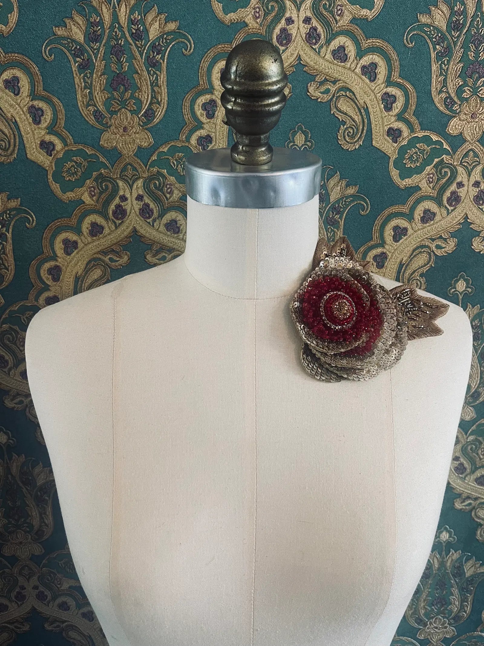 Broochella tonina tesorina gold and red hand beaded flower brooch. Pictured on a mannequin. The background is green and gold damask wallpaper