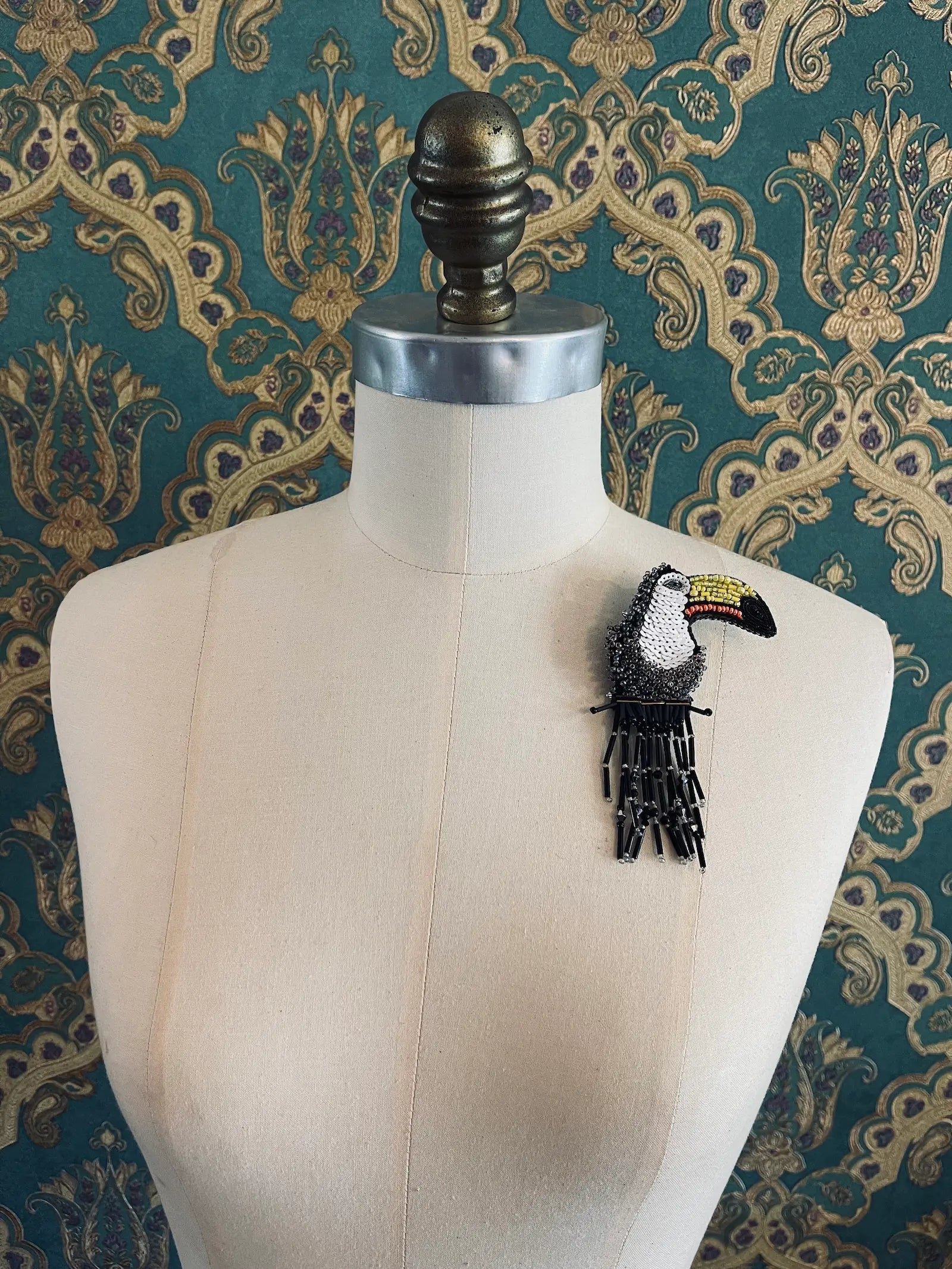 Broochella Toucan hand beaded bird brooch in beads and sequins. Pictured on a mannequin. The background is green and gold damask wallpaper.