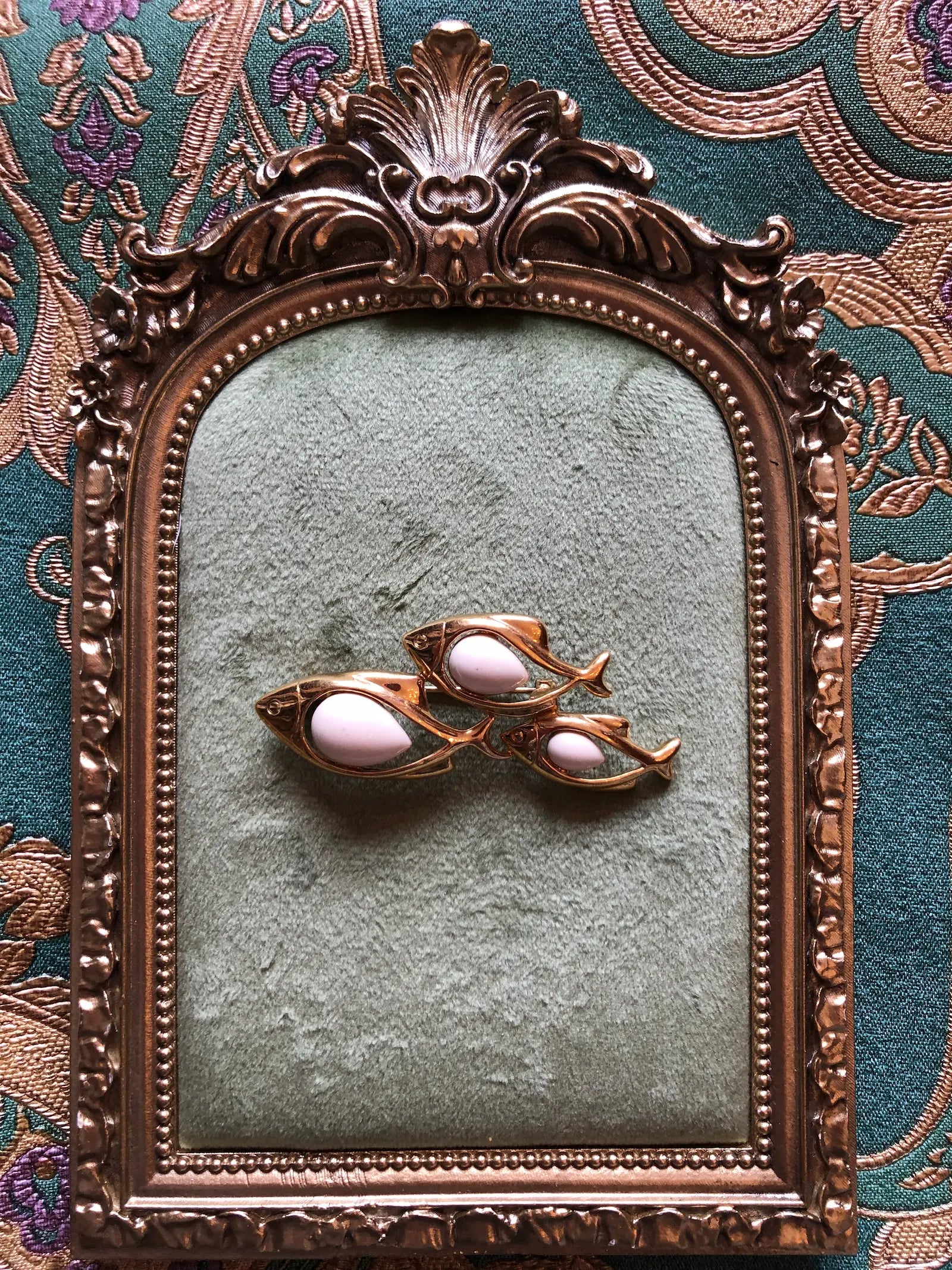 Broochella Tre Pesci vintage gold-tone brooch featuring three swimming fish with smooth ivory enamel bodies, displayed on a textured sage green surface inside an ornate vintage-style bronze mirror frame.