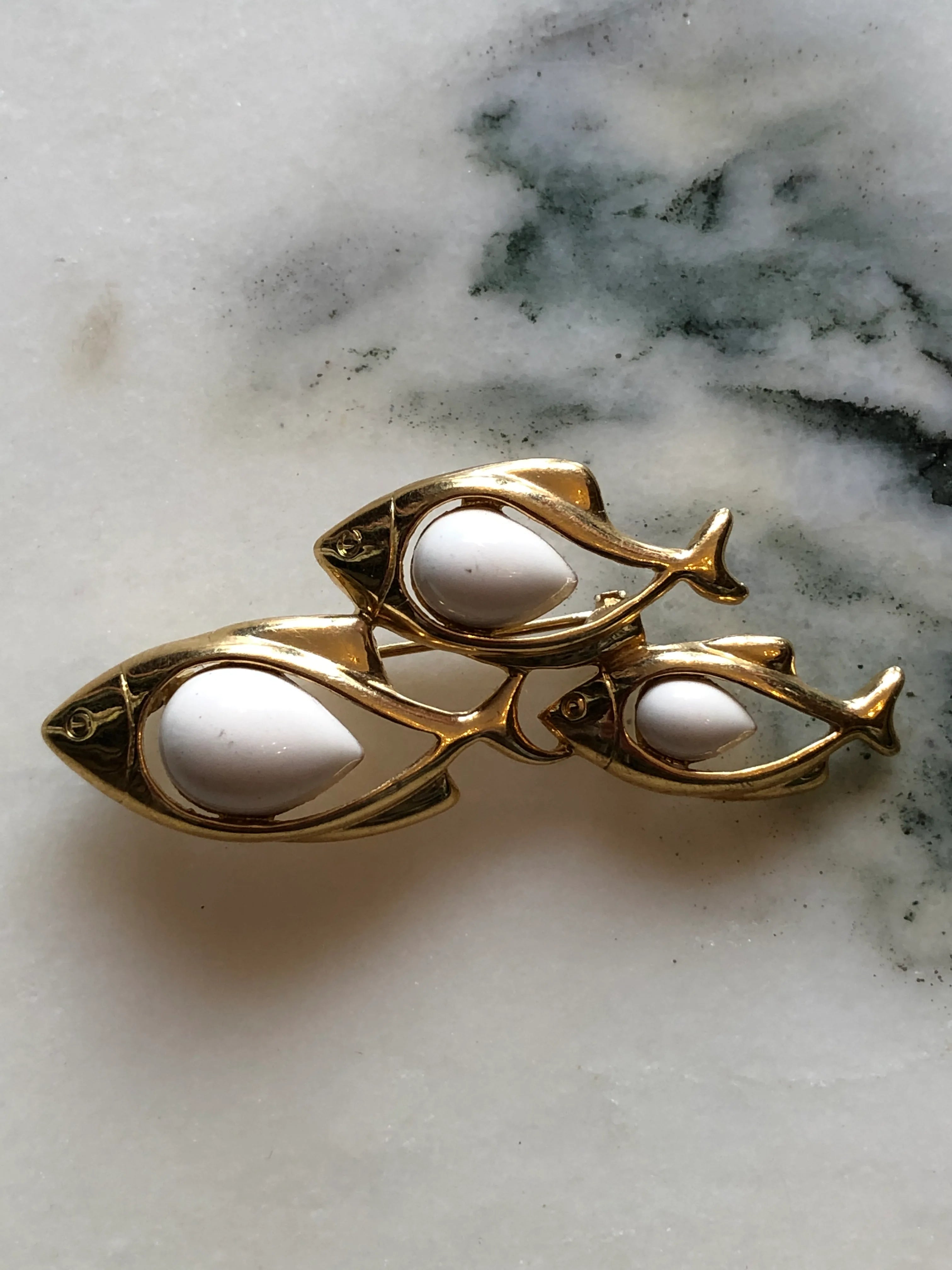 Broochella Tre Pesci vintage gold-tone brooch featuring three swimming fish with smooth ivory enamel bodies. The background is white marble.