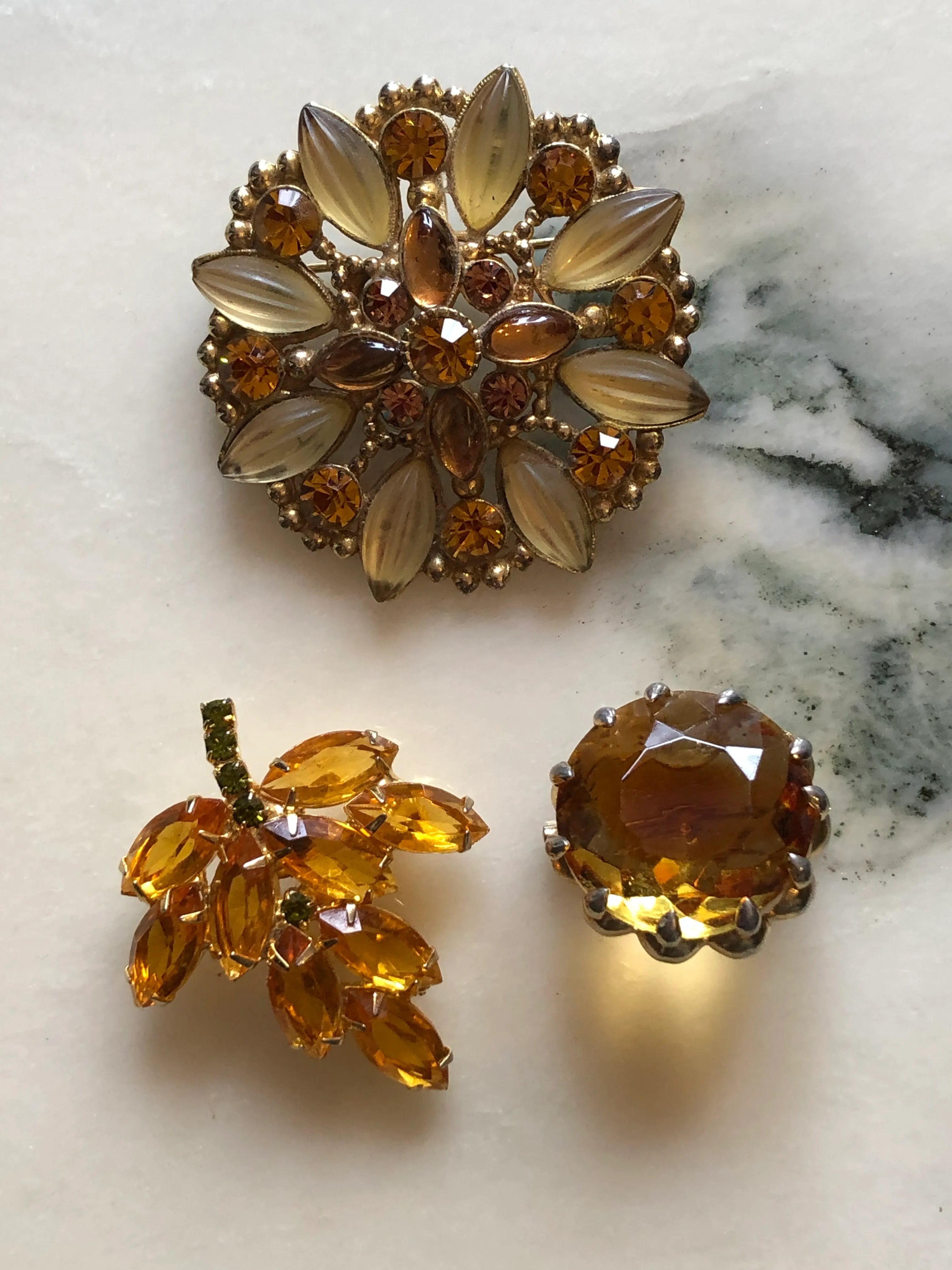 Broochella Tris d’Ambra set of three vintage amber crystal brooches, including a stamped Goldcrest floral brooch. The background is white marble