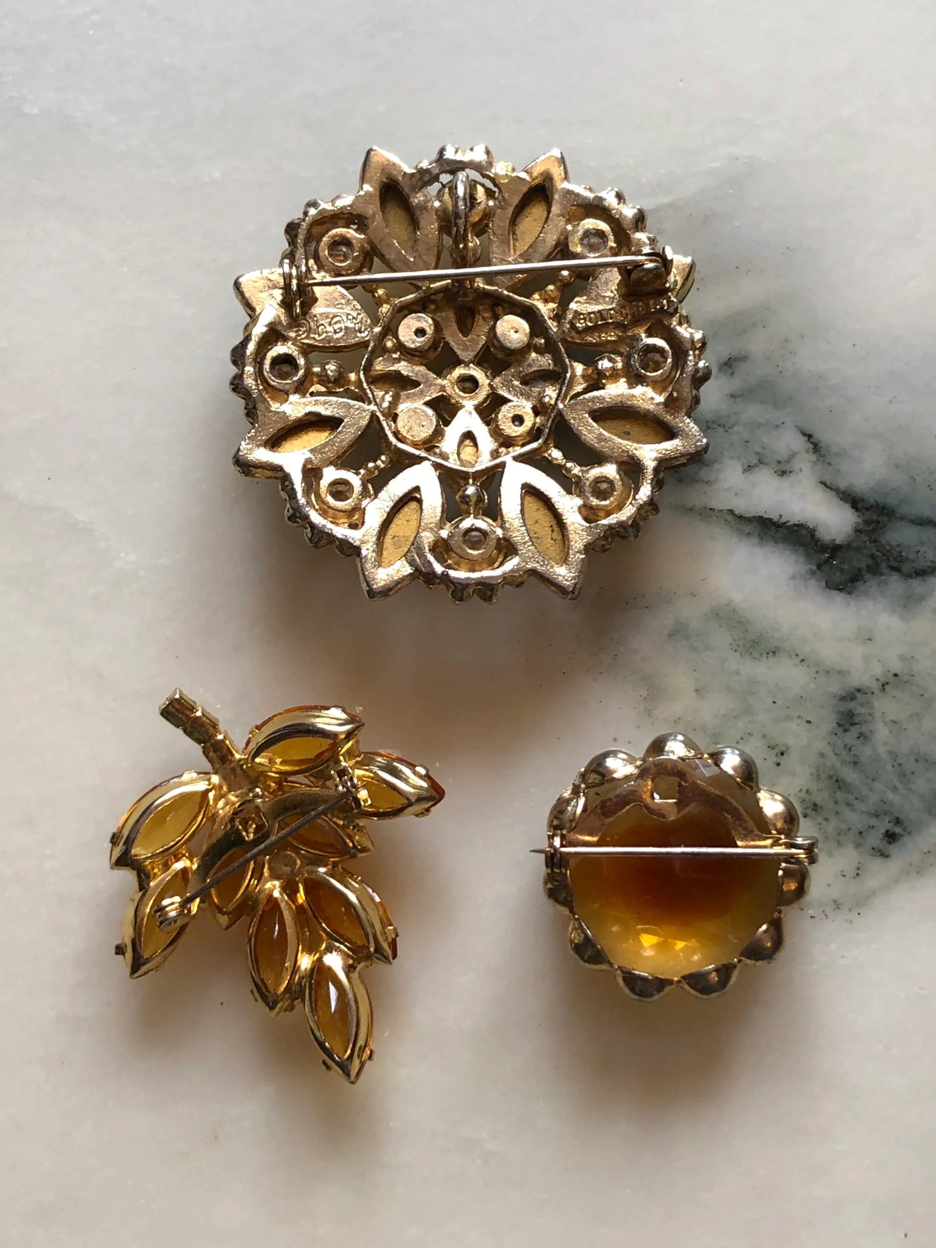 The back view of Broochella Tris d’Ambra set of three vintage amber crystal brooches, including a stamped Goldcrest floral brooch. The background is white marble