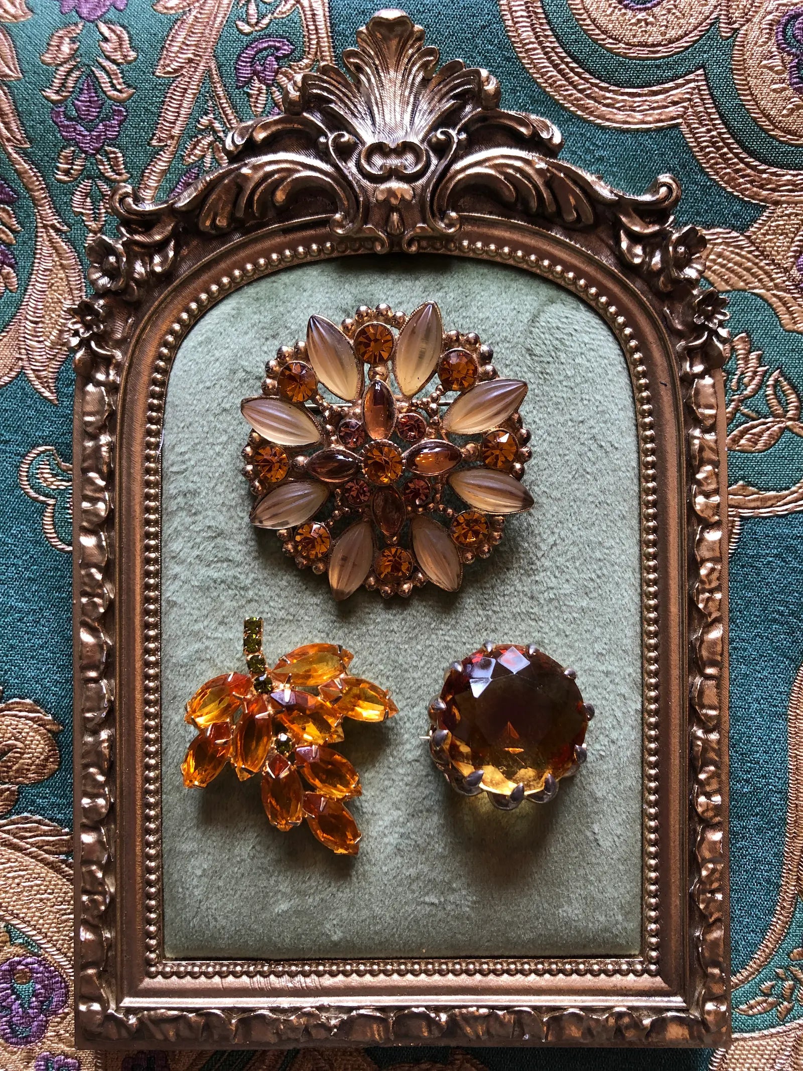 Broochella Tris d’Ambra set of three vintage amber crystal brooches, including a stamped Goldcrest floral brooch. Displayed inside an ornate antique gold frame against a teal patterned fabric background.