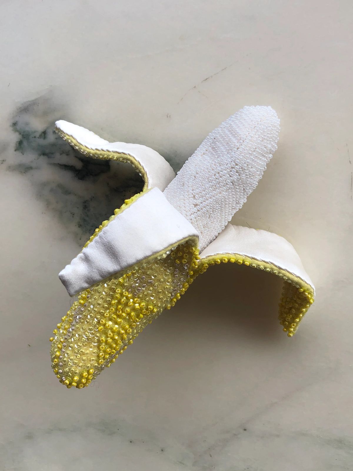 Broochella Tropicana Banana Brooch Pin, a playful statement banana design hand-embellished with white, yellow and clear beads and sequins, photographed on white marble with soft grey-green veining.