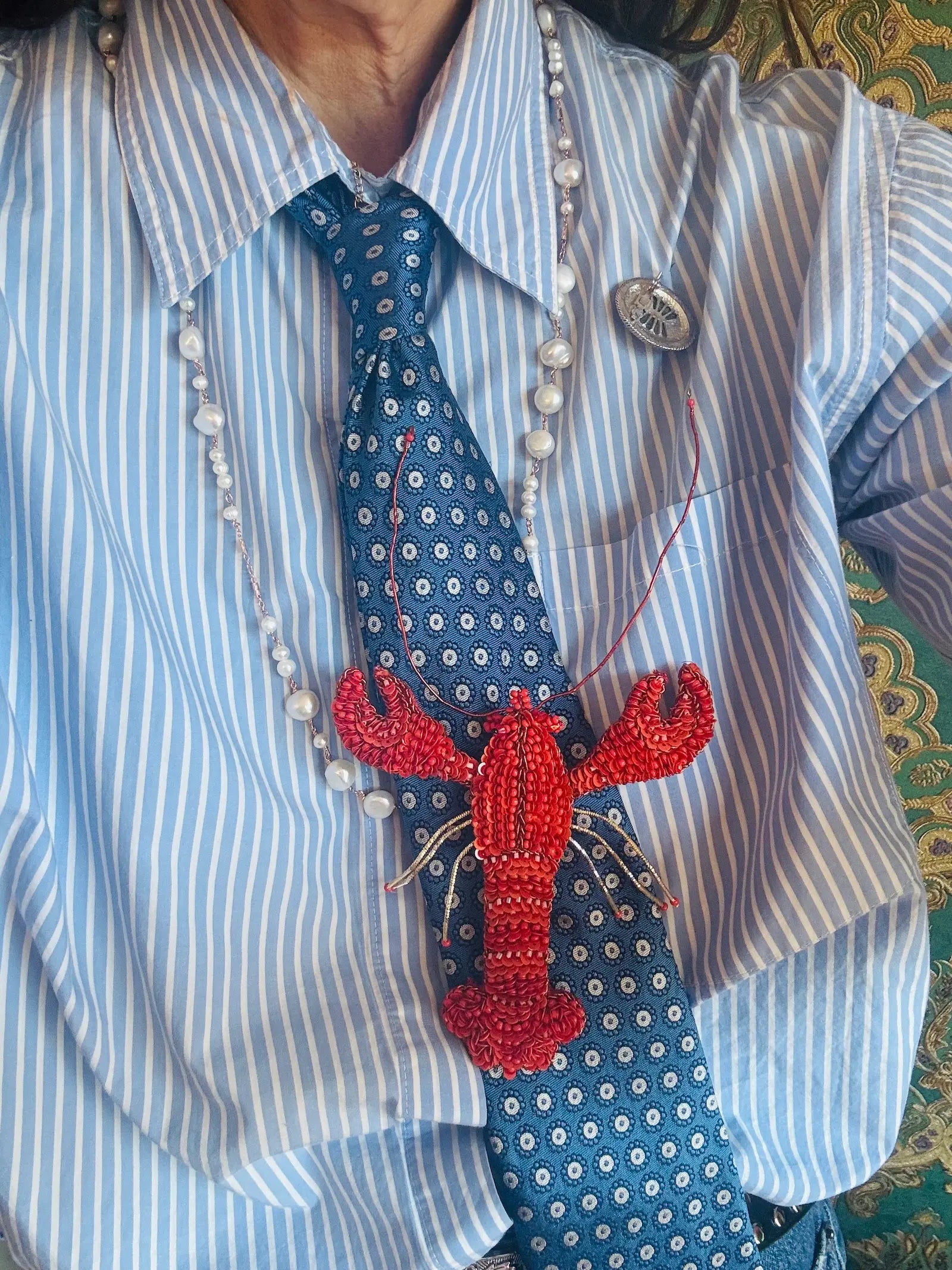 A close-up of a person wearing a light blue and white striped button-down shirt, a blue patterned necktie, a beaded white pearl necklace, and a Broochella-Tropicana red beaded lobster brooch pinned at the centre of the chest.