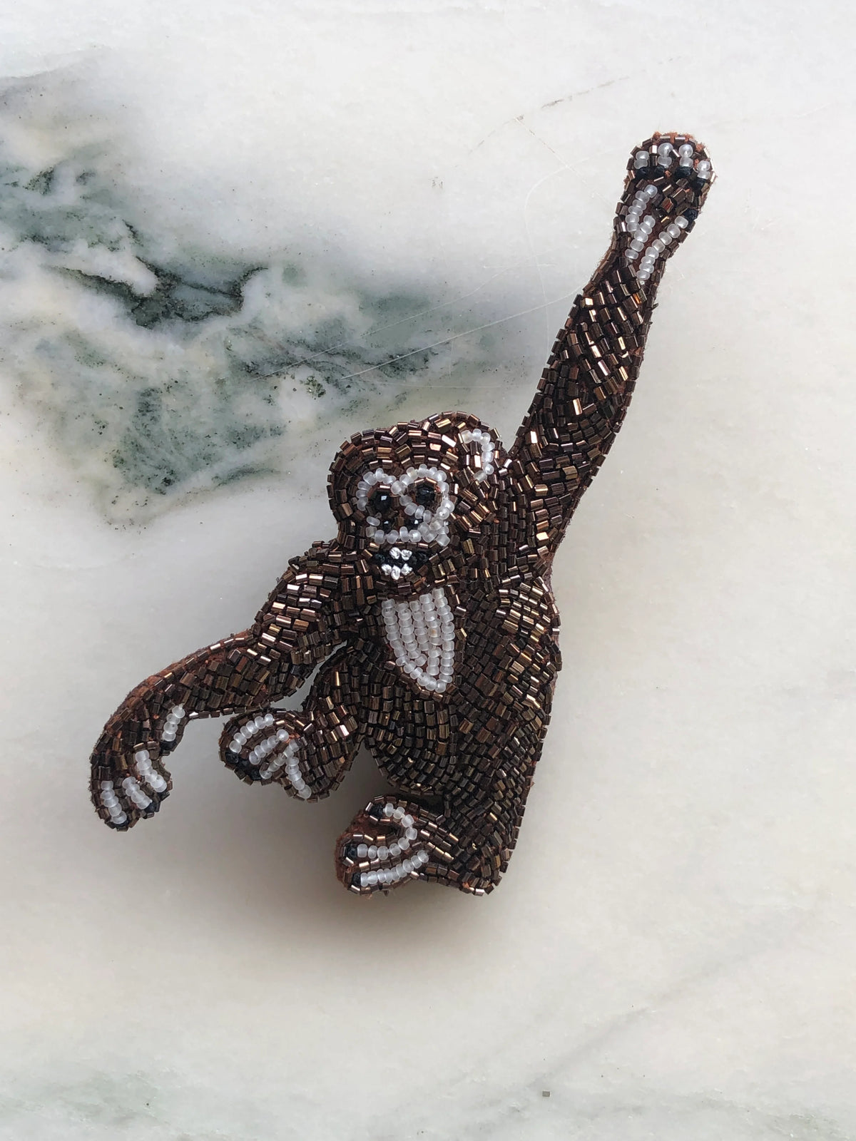 Broochella Tropicana Monkey Brooch Pin, a playful statement monkey design intricately hand-beaded in bronze, brown, black and white beads, photographed on white marble with soft grey-green veining.