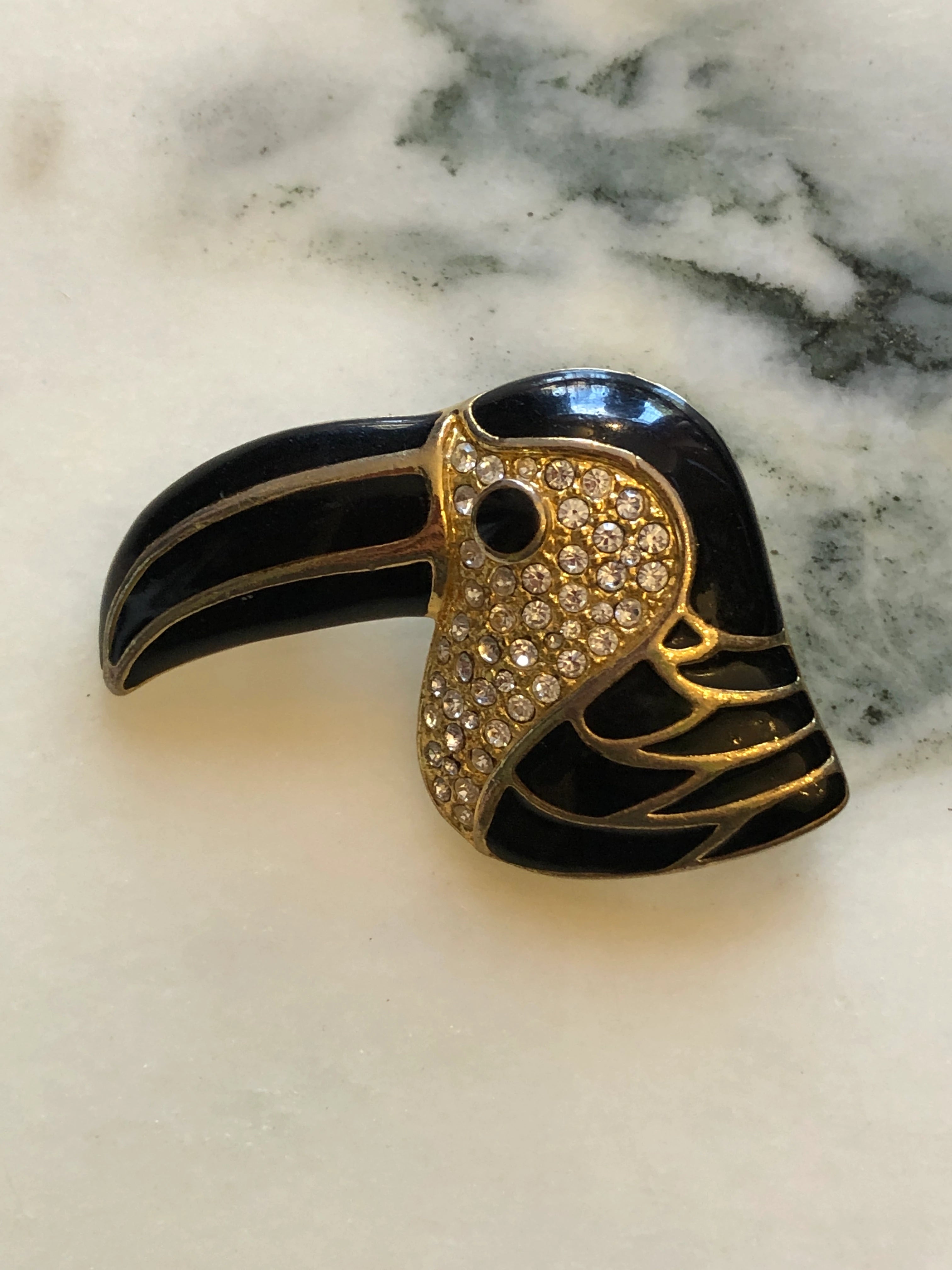 A vintage Broochella Toucano Nero  toucan head brooch featuring black enamel, gold-tone metal, and clear rhinestones on a light marble surface.