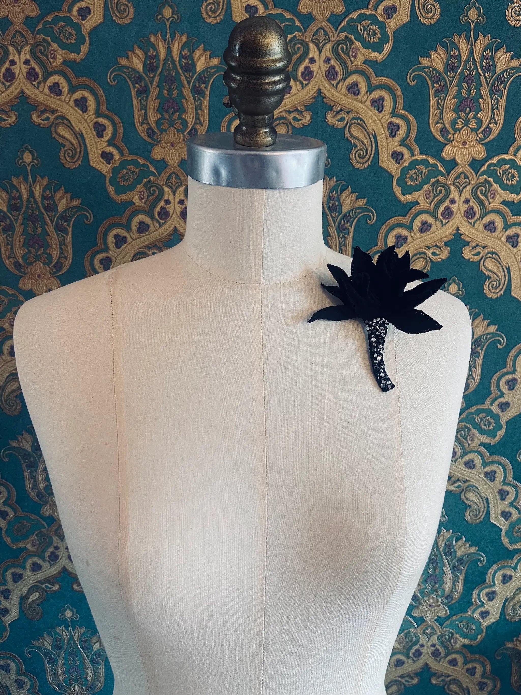 Broochella Tulipano small handmade black fabric and crystal flower brooch. Pictured on a mannequin. The background is green and gold damask wallpaper