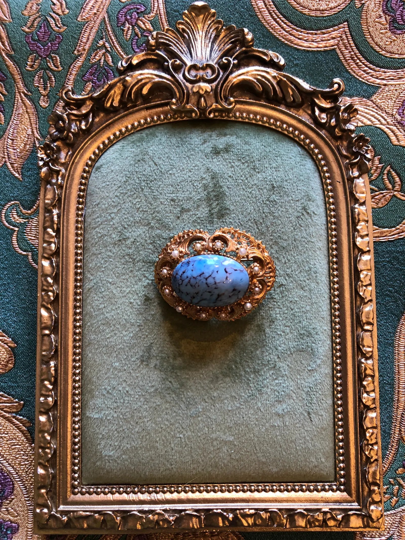 Broochella Turchese vintage gold-tone brooch with a turquoise-blue marbled cabochon, ornate filigree and faux pearl accents. Pictured on a green velvet tray with an ornate gold frame. The background is green and gold damask wallpaper