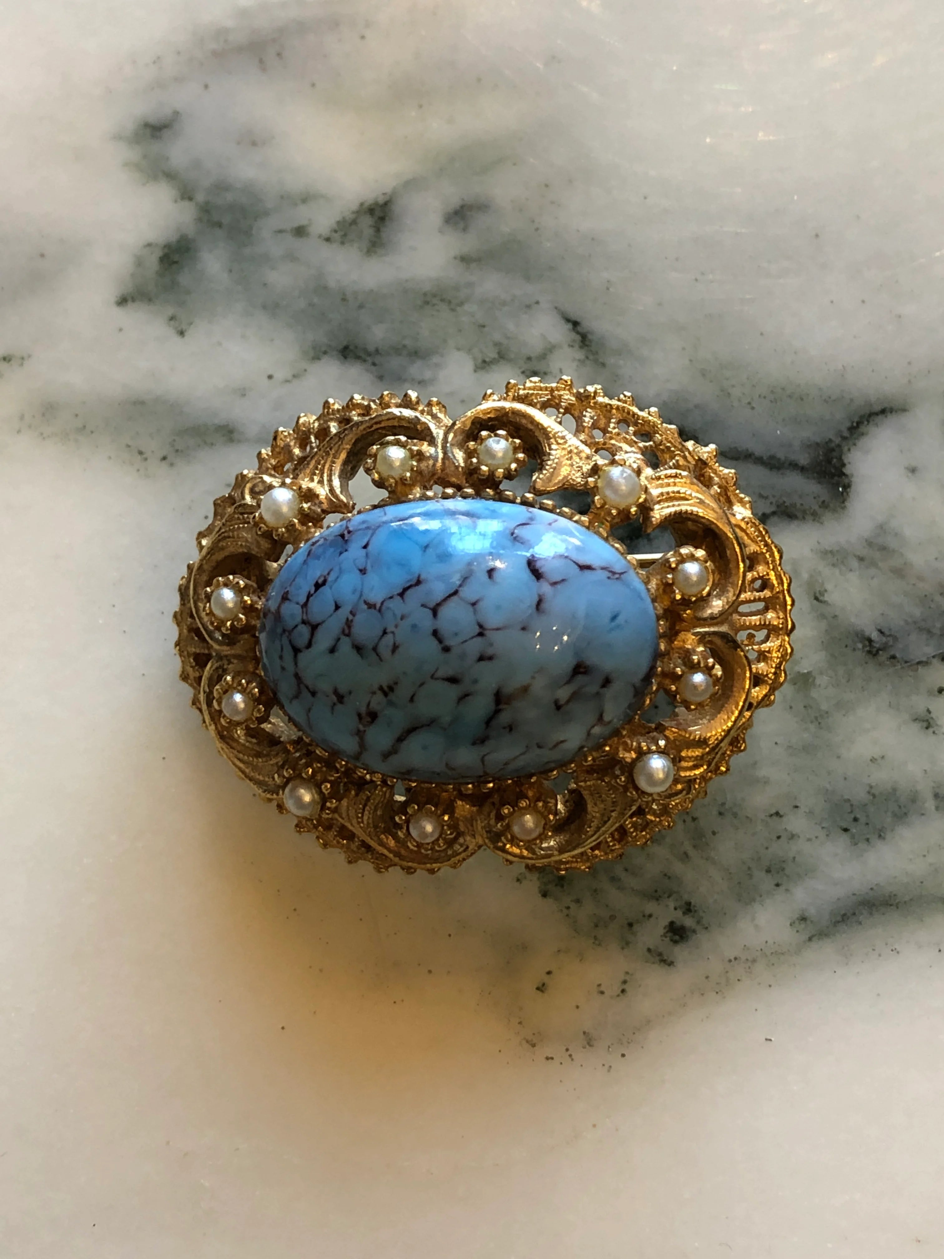 Broochella Turchese vintage gold-tone brooch with a turquoise-blue marbled cabochon, ornate filigree and faux pearl accents The background is white marble
