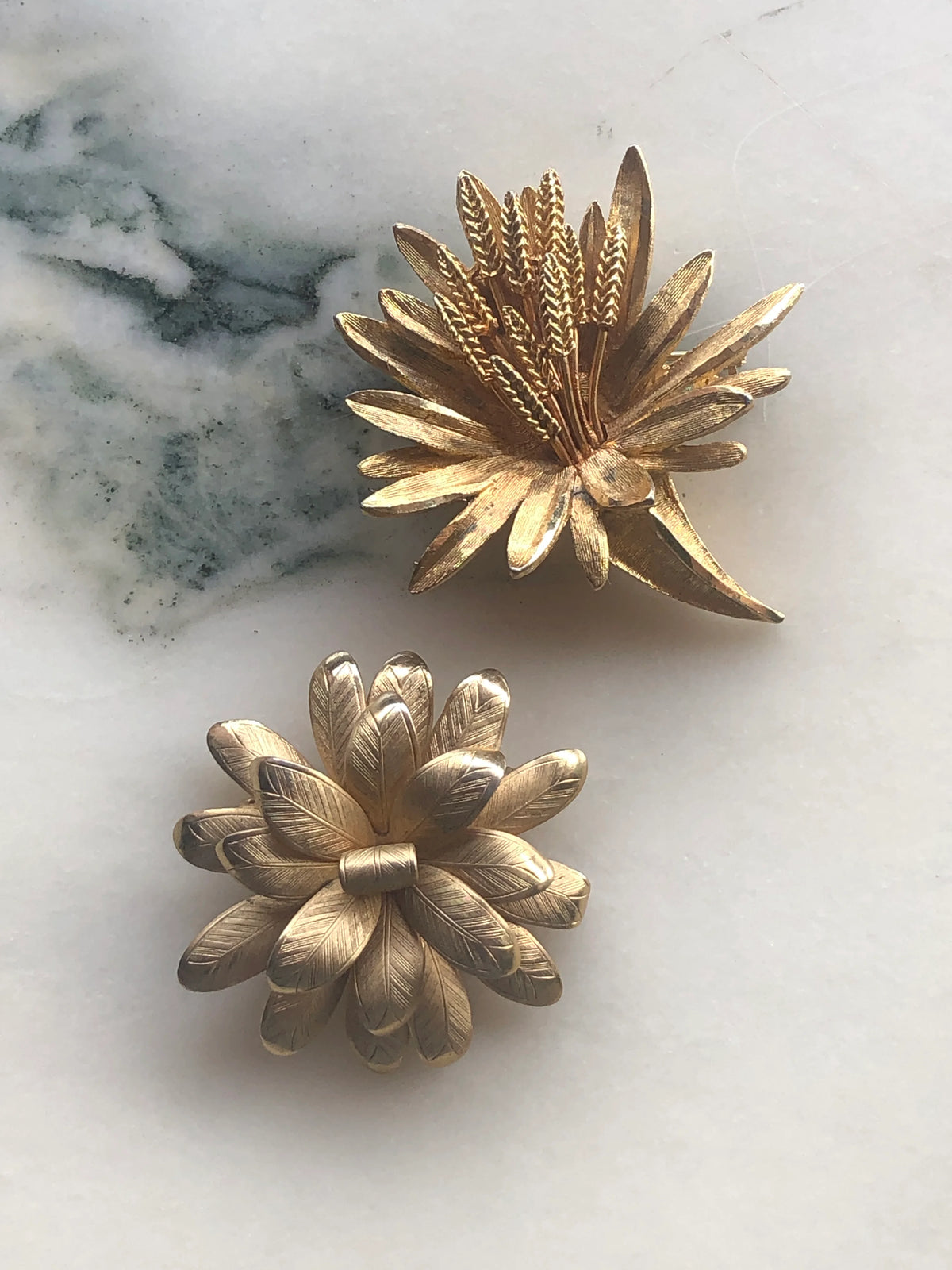 Broochella Two Golden Blooms Vintage Brooch Set featuring two different gold-tone floral designs with layered textured petals and botanical detailing, photographed on white marble with soft grey-green veining.