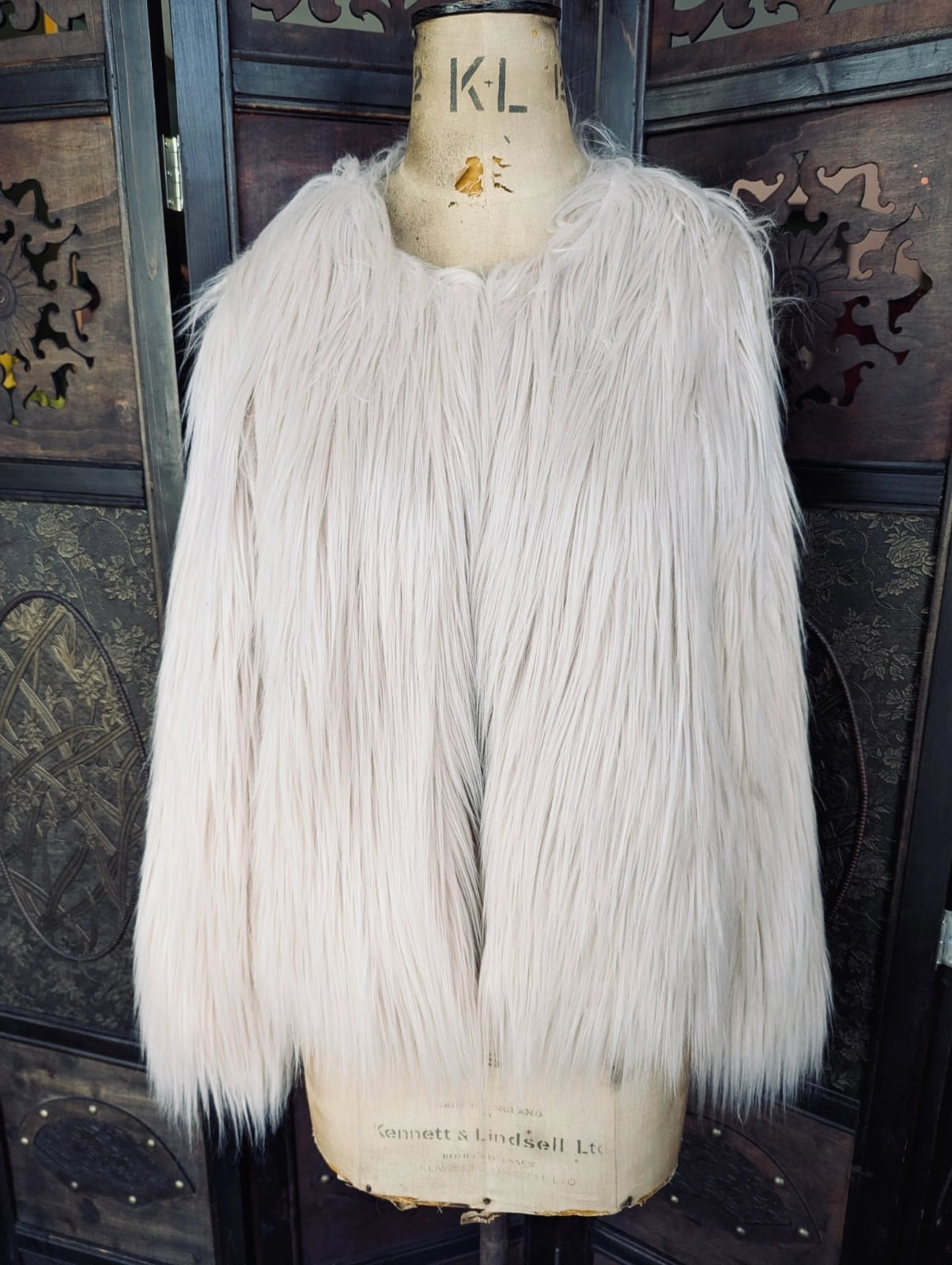 Broochella Unreal Fur Faux fur coat in beige. It is pictured on a vintage mannequin. The background is an ornate, dark wood room divider. 