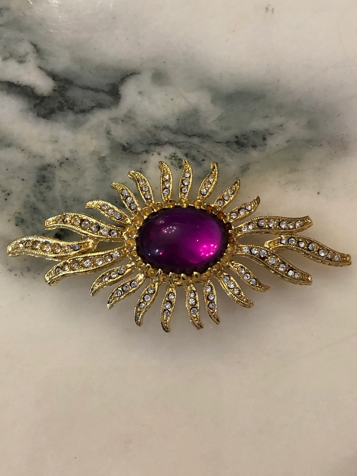 Broochella Valentina Vintage Purple Sunburst Brooch featuring a deep-purple oval cabochon surrounded by gold-tone crystal-set rays on a cream and grey marble background.