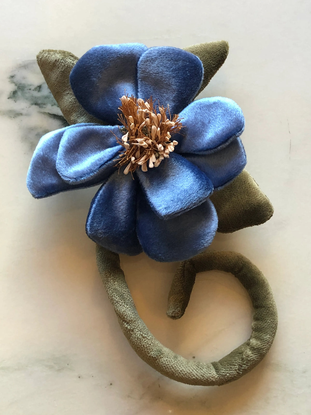 A Broochella Velluto handmade fabric flower brooch with blue velvet petals, olive green velvet leaves and stem, and a metallic bronze center, displayed on a light marble surface.