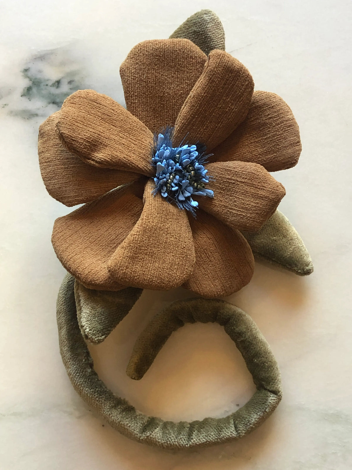 A Broochella Men's Velluto handmade velvet fabric flower brooch featuring large brown petals, olive green leaves, and a blue flower centre,  displayed on a light marble surface.